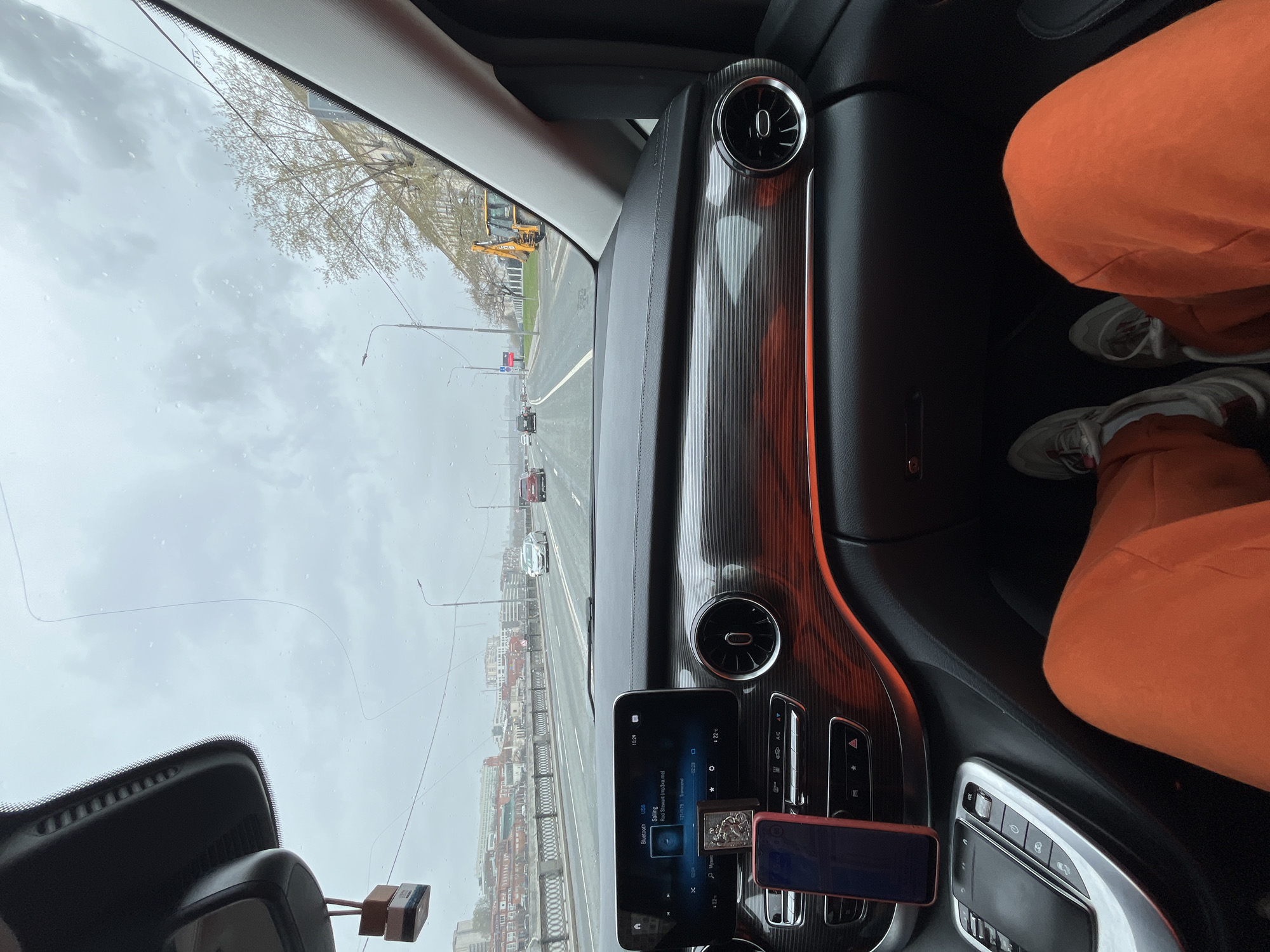 The image shows the view from inside a car driving on a bridge in Moscow, Russia. The weather is overcast and it is raining. Multiple buildings can be seen on the left. Traffic is light with multiple vehicles travelling on the road, in the same direction as the car. A construction vehicle is visible in the distance. Inside the car, there is the interior trim of the vehicle and part of the navigation system is visible, playing a song by Rod Stewart. On the right is part of a person wearing orange clothing and white tennis shoes.