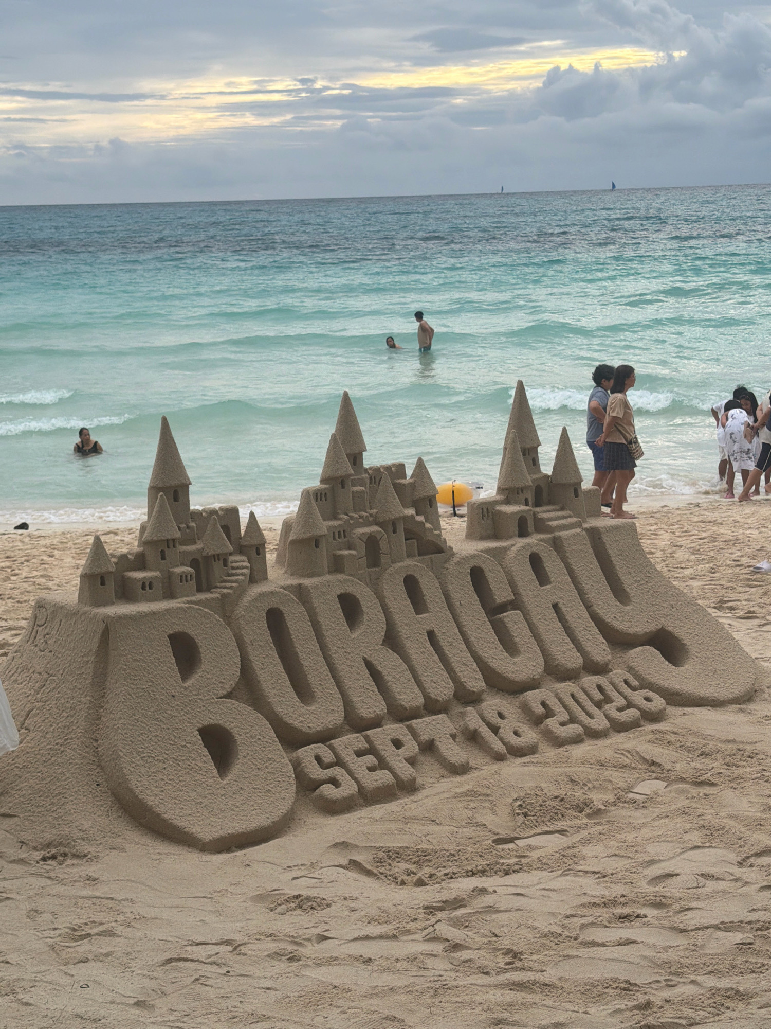 The image depicts a sandcastle on the beach of Boracay, a popular tourist island in Malay, Aklan, Philippines. The sandcastle is elaborately constructed, forming the word "BORACAY" in large, three-dimensional letters. Below the name, the date "SEPT 18 2025" is also sculpted into the sand. Behind the sandcastle, the turquoise waters of the ocean meet the sandy shore. Several people are visible in the water and on the beach, some swimming or wading, while others stand in small groups. The sky above is overcast with clouds, hinting at a potential change in weather. The overall scene captures a moment of leisure and artistic expression on a tropical beach.