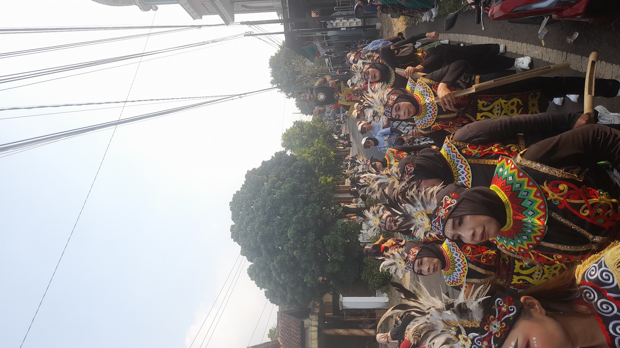 In Lemahabang, Indonesia, a group of people in traditional attire are participating in a procession along a street. The individuals in the foreground are women adorned with elaborate headdresses featuring feathers and decorative patterns, and they wear dark tops with brightly colored, intricately embroidered collars. They are holding wooden poles. Behind them, more participants are visible, some carrying a large effigy of a man. The procession is taking place on a paved road lined with houses and trees. Utility wires crisscross the sky above. The scene appears to be a cultural or religious event, possibly a parade or festival, occurring during daylight hours.
