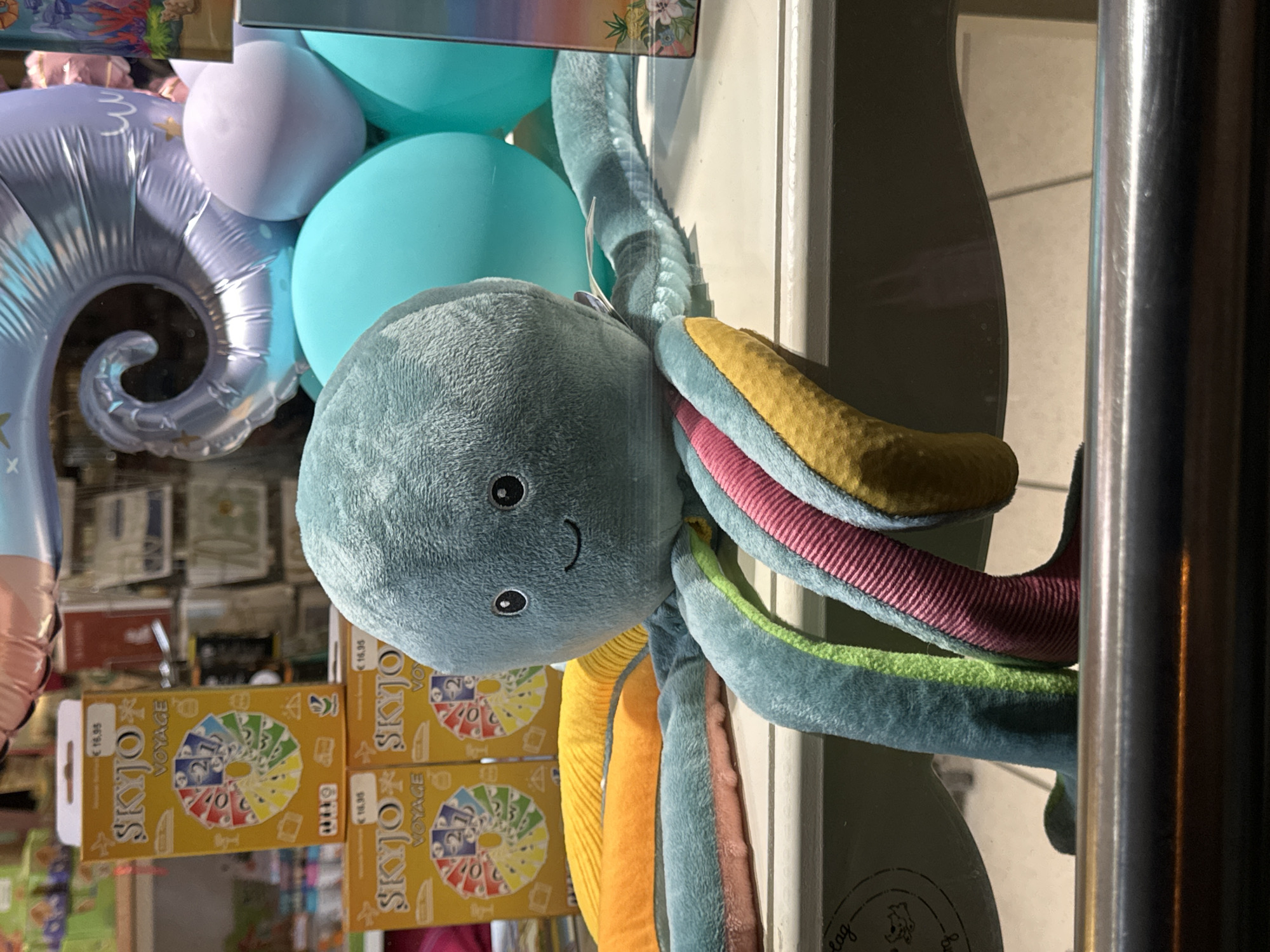 The image shows a plush octopus toy with a teal-colored body and multi-colored tentacles. The octopus is positioned in the foreground, resting on a white shelf. Behind the octopus, there are several boxes of a game called "SKYJO VOYAGE," as well as some large, round balloons in shades of teal and lavender. The overall scene appears to be a retail display, likely in a toy store or a gift shop. The specific location in Krefeld, Germany, is not determinable from the visual information provided in the image.