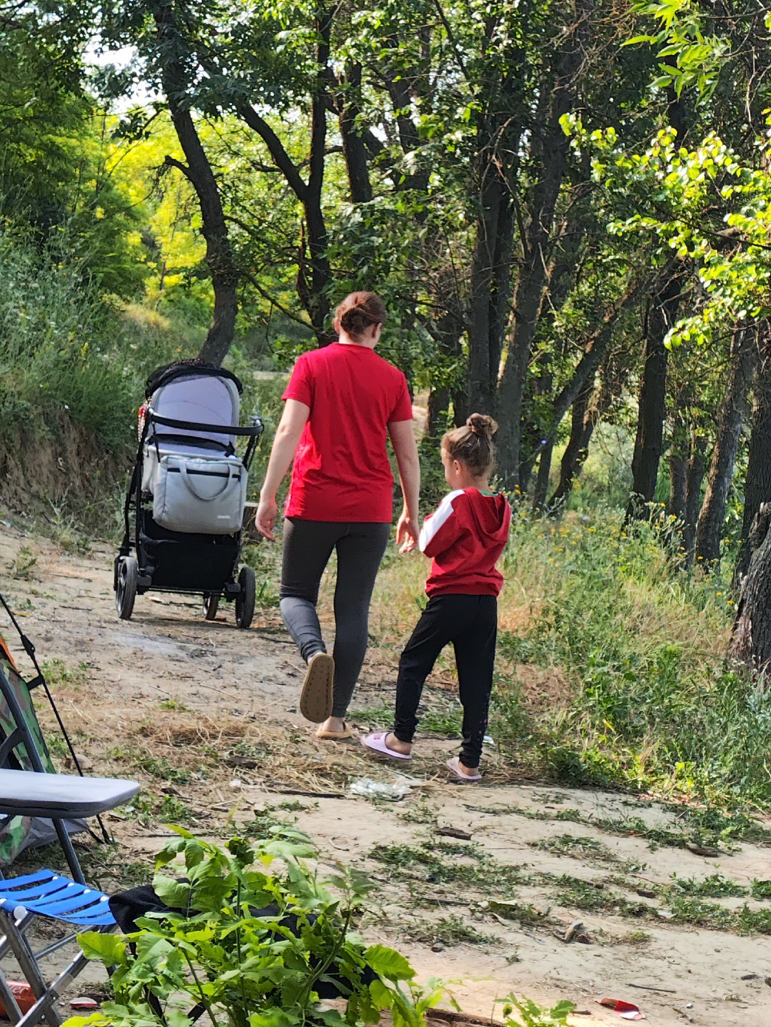 In Găleștii Noi, Moldova, a woman in a red t-shirt and gray leggings walks away from the camera, pushing a stroller with a gray bag attached. A young girl in a red hooded sweatshirt with white stripes and black pants walks slightly ahead of the woman, also facing away from the camera. They are on a dirt path surrounded by lush green trees and vegetation. In the foreground, to the left, are parts of a blue folding chair and a tent. The overall scene depicts a family outing in a natural, wooded area.