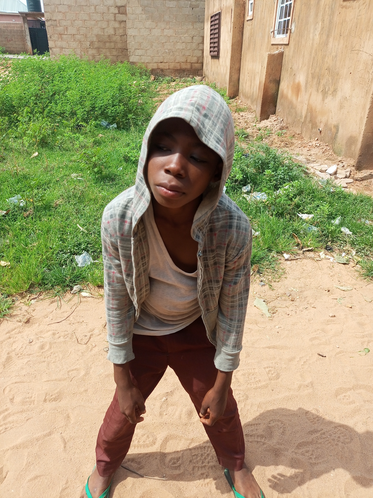 A young person, wearing a plaid hoodie over a light-colored t-shirt and maroon pants, stands in a sandy area outdoors. The person's gaze is directed downwards, and their posture suggests a moment of rest or contemplation. Green grass and some scattered debris are visible behind them, bordering a light brown building constructed of brick. The building has windows and architectural details suggesting a weathered appearance. The ground is sandy and uneven, with footprints and shadows cast by the bright sunlight, indicating it is daytime. The setting appears to be a rural or semi-urban area in Tempure, Nigeria, with the building and vegetation providing context.