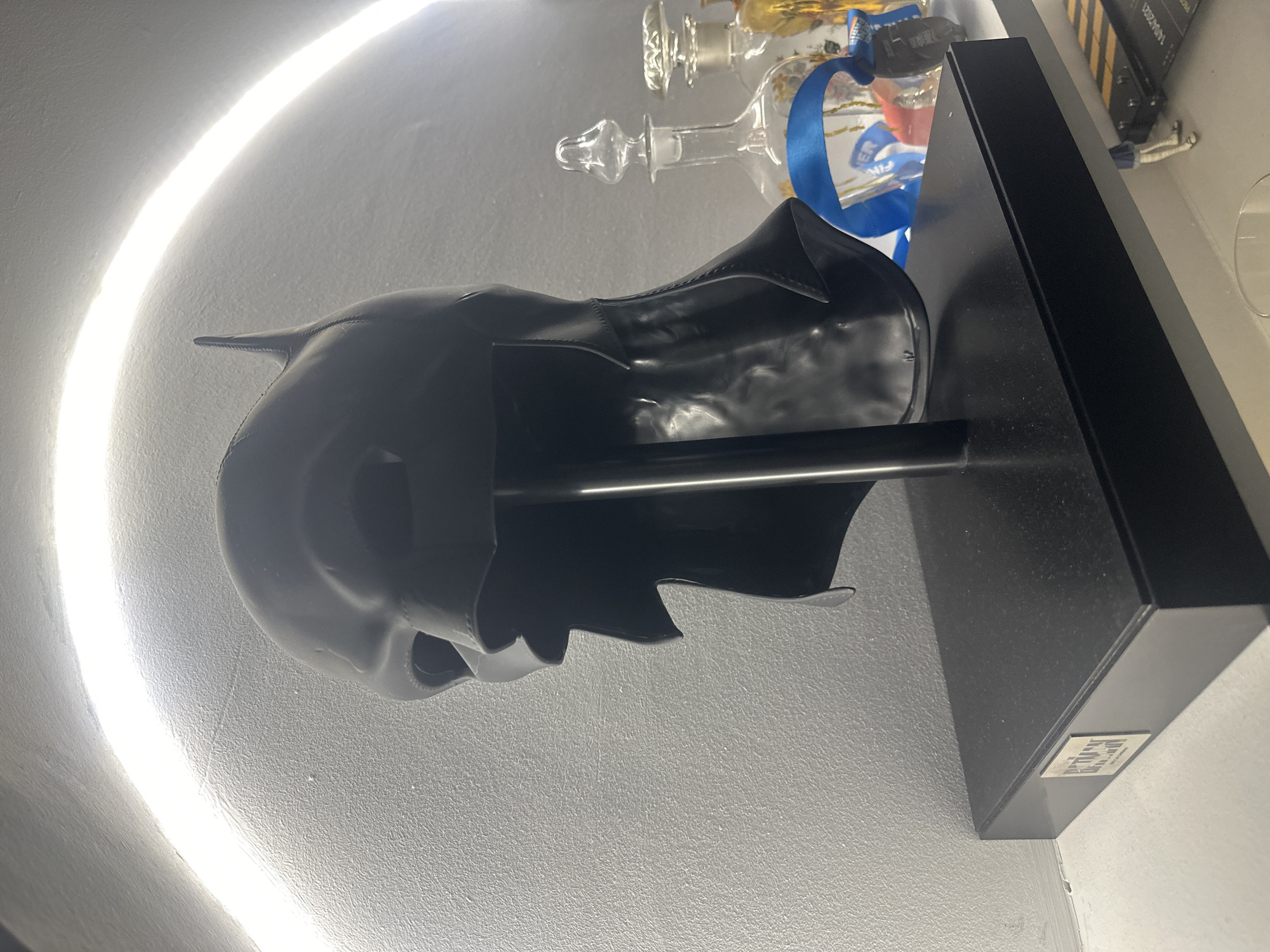 The image shows a Batman mask displayed on a black stand in front of a white wall. The mask is black and appears to be made of a smooth material. A glass bottle with a blue ribbon around it is visible in the background. The location is in Clichy, France.
