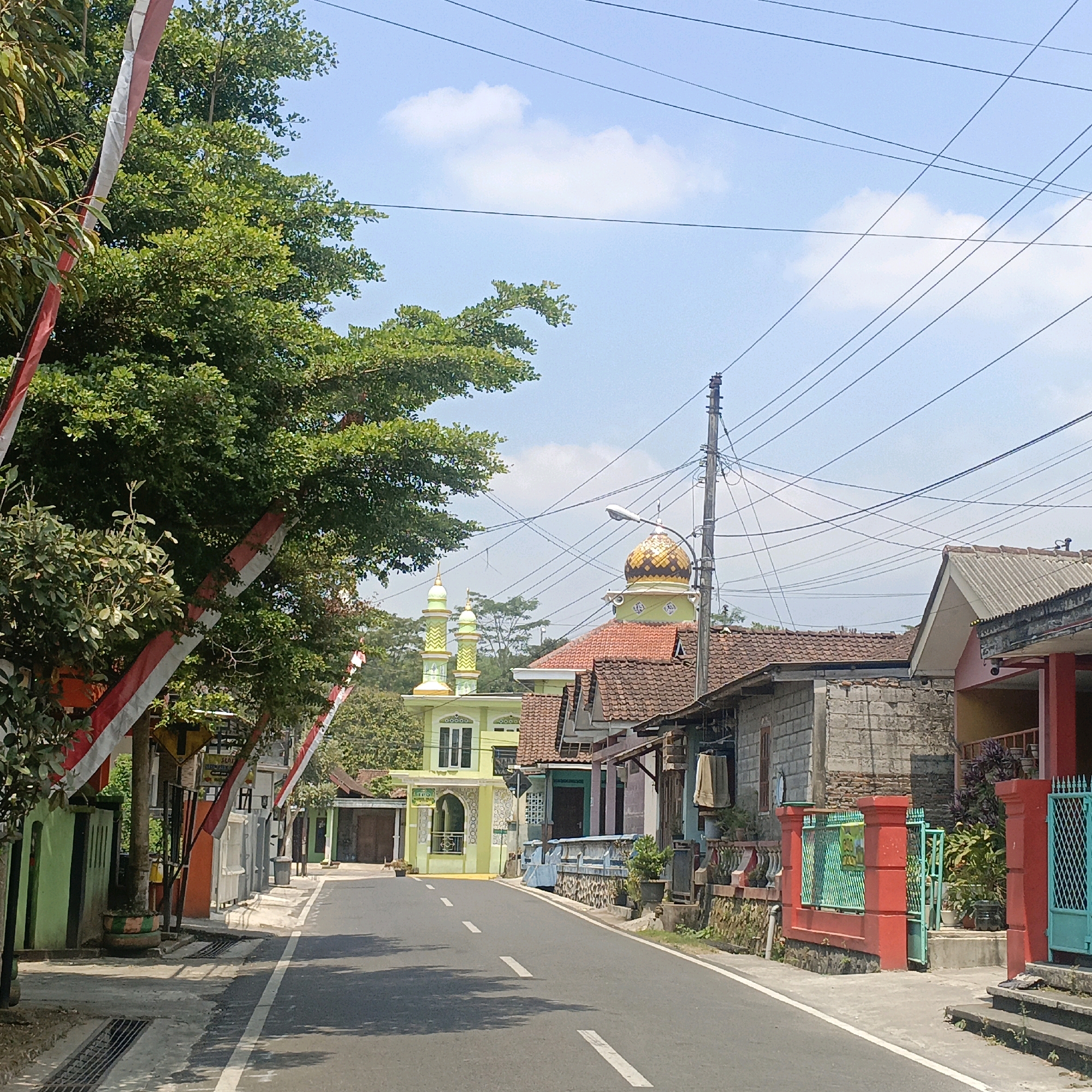 The image captures a street scene in Salatiga, Indonesia, likely in a residential or mixed-use area, featuring a mosque and several houses. The road, paved with asphalt, has white dashed lines indicating lanes. On the left side of the road, a large, leafy tree is adorned with red and white Indonesian flags. Beyond the tree, there are houses with green and grey facades, and a sidewalk with a drainage grate. Further down the street on the left is a light green building with a minaret, appearing to be a mosque or a similar religious structure, decorated with intricate patterns and a golden dome.
On the right side of the road, a series of houses are visible. The first is a two-story building with a tiled roof, followed by a single-story structure with a light-colored brick facade. This is adjacent to a house with a red gate and a light blue gate further down. The houses display various architectural styles and colors, including red, grey, and light blue accents. A telephone pole with numerous wires is prominently positioned in the middle-right of the frame. The sky is a clear blue with a few scattered white clouds. The overall impression is a typical street in a tropical climate, possibly decorated for a national holiday. There are no people visible in the image.