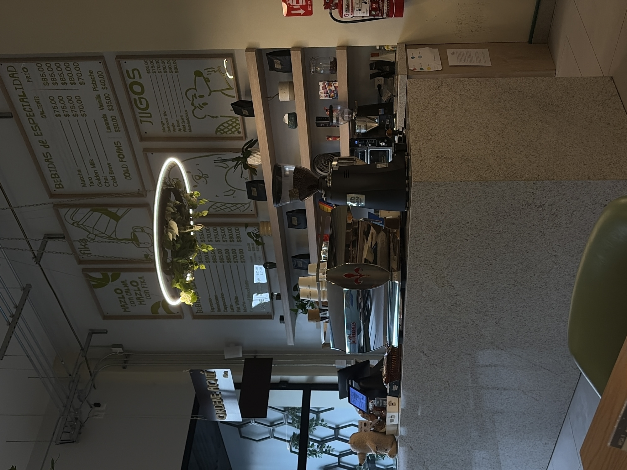 This image shows the interior of a cafe or coffee shop, possibly in Puebla, Mexico, given the Spanish signage. A counter with a stone-like finish dominates the view, behind which is coffee-making equipment, including an espresso machine and a grinder. Shelves behind the equipment hold supplies and decorative items. Signage above displays menu items and prices for "Bebidas de Especialidad" (specialty drinks) and "Jugos" (juices). A circular light fixture surrounds greenery. A sign that reads "ORDENE AQUI" indicating an ordering point is visible. The environment suggests a modern cafe setting with an emphasis on coffee and juice offerings.