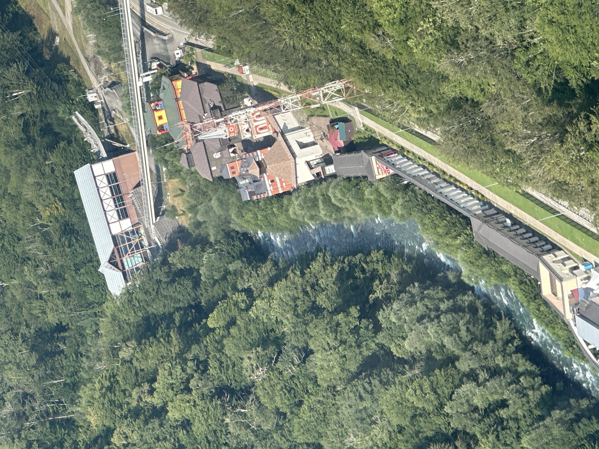 This is an aerial, high-angle, full shot of a resort area in Estosadok, Russia, nestled within a dense, green forest. A turbulent river flows through the center of the image, bordered by a well-maintained pathway. Along the pathway, there are several buildings with distinct architectural styles.
To the left of the river, a long, multi-story building with a grey roof and white accents runs parallel to the pathway. Further up the river and to the right, a complex of buildings with colorful roofs (yellow, green, and red) and ornate details stands prominently. One building features a red tower with white accents, resembling a clock tower or church steeple, topped with a spire. A large transmission tower, painted with red and white sections, is situated near these colorful buildings.
Above the colorful buildings, a modern, multi-level structure with a white roof and glass-paneled walls is visible, connected by elevated walkways and bridges to higher ground. A road runs alongside some of these structures, with a white vehicle and a white bus visible. The surrounding environment is dominated by lush, green trees, indicating a natural, forested setting. The overall impression is of a developed tourist destination integrated into a natural landscape.
