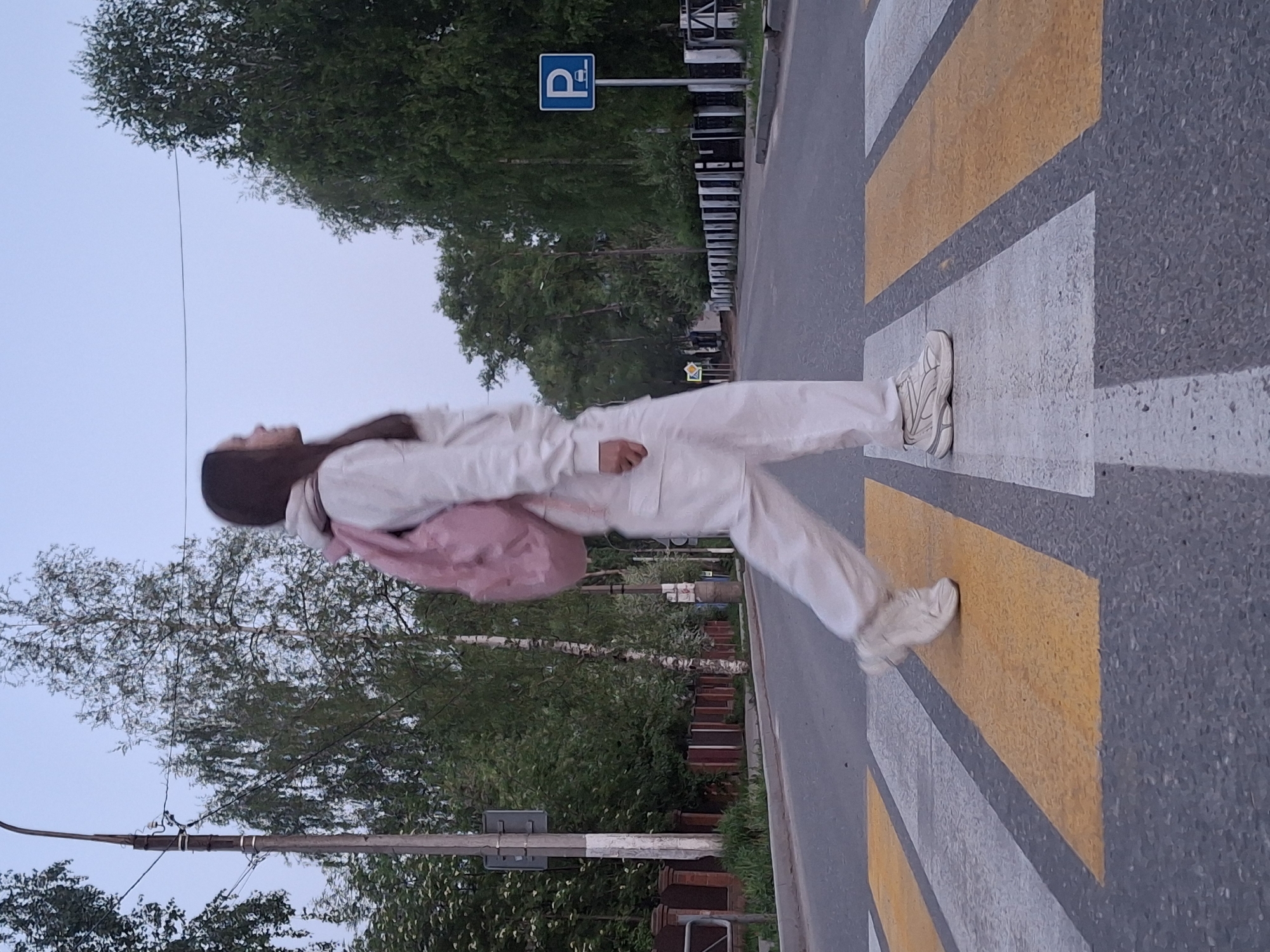 A woman wearing a white tracksuit and a pink backpack is walking across a pedestrian crosswalk in the city of Chaikovsky, Russia. The crosswalk is marked with alternating yellow and white stripes on an asphalt road. A parking sign is visible in the background among green trees and other vegetation. A lamppost stands to the left.