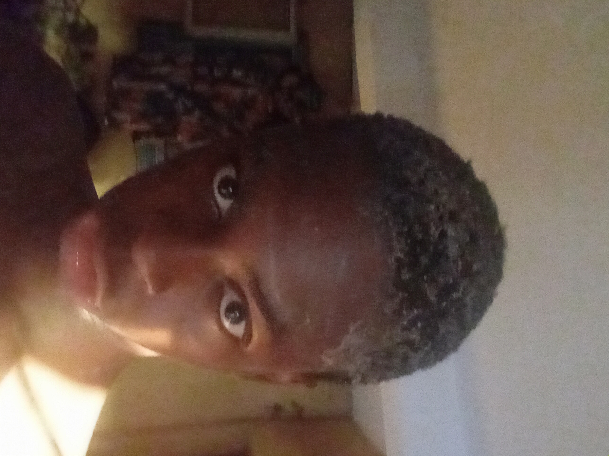 A close-up, tilted portrait shows a person with dark skin and short, textured black hair. The background is blurry, but suggests an interior space, possibly in a residential setting, with furniture or items on a shelf. The image is taken indoors. The city of Nkpor, Nigeria is not discernible in this image.