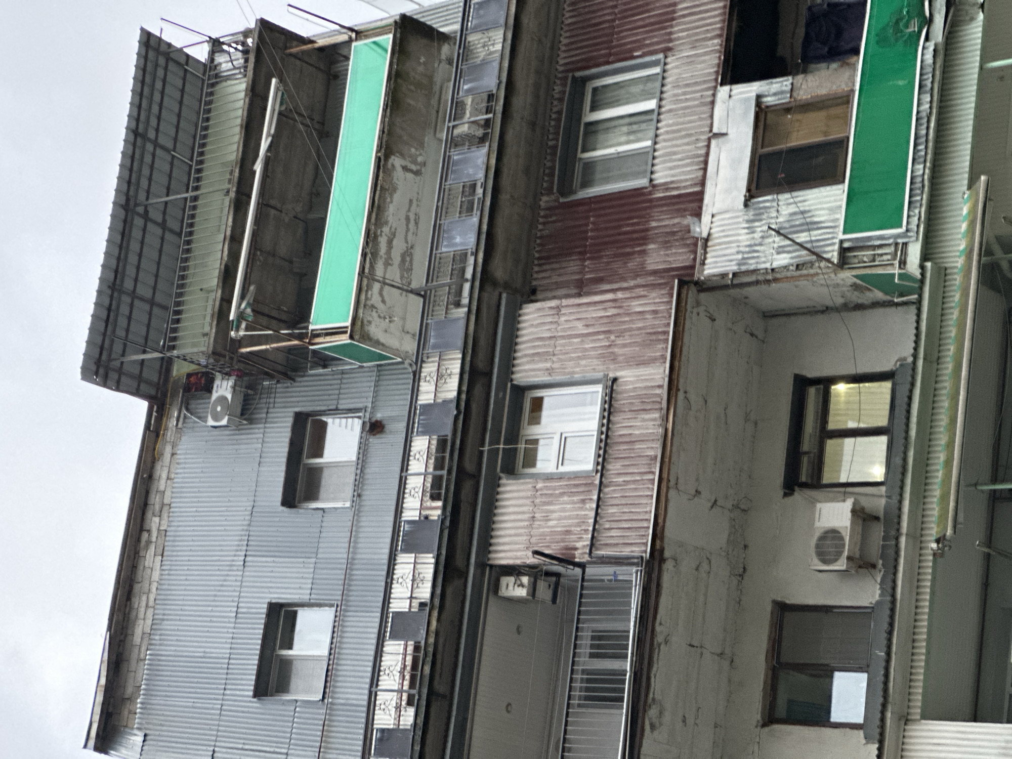 The image shows a narrow, decaying building exterior in what appears to be a dense urban environment. The building has several stories, with each level exhibiting makeshift modifications and additions. There is corrugated metal siding, with multiple windows of varying sizes and styles. The building features several protruding structures covered with green fabric, likely for shade or privacy. The overall impression is one of age, disrepair, and improvised construction. The specific location in Batumi, Georgia, cannot be determined from this image alone.