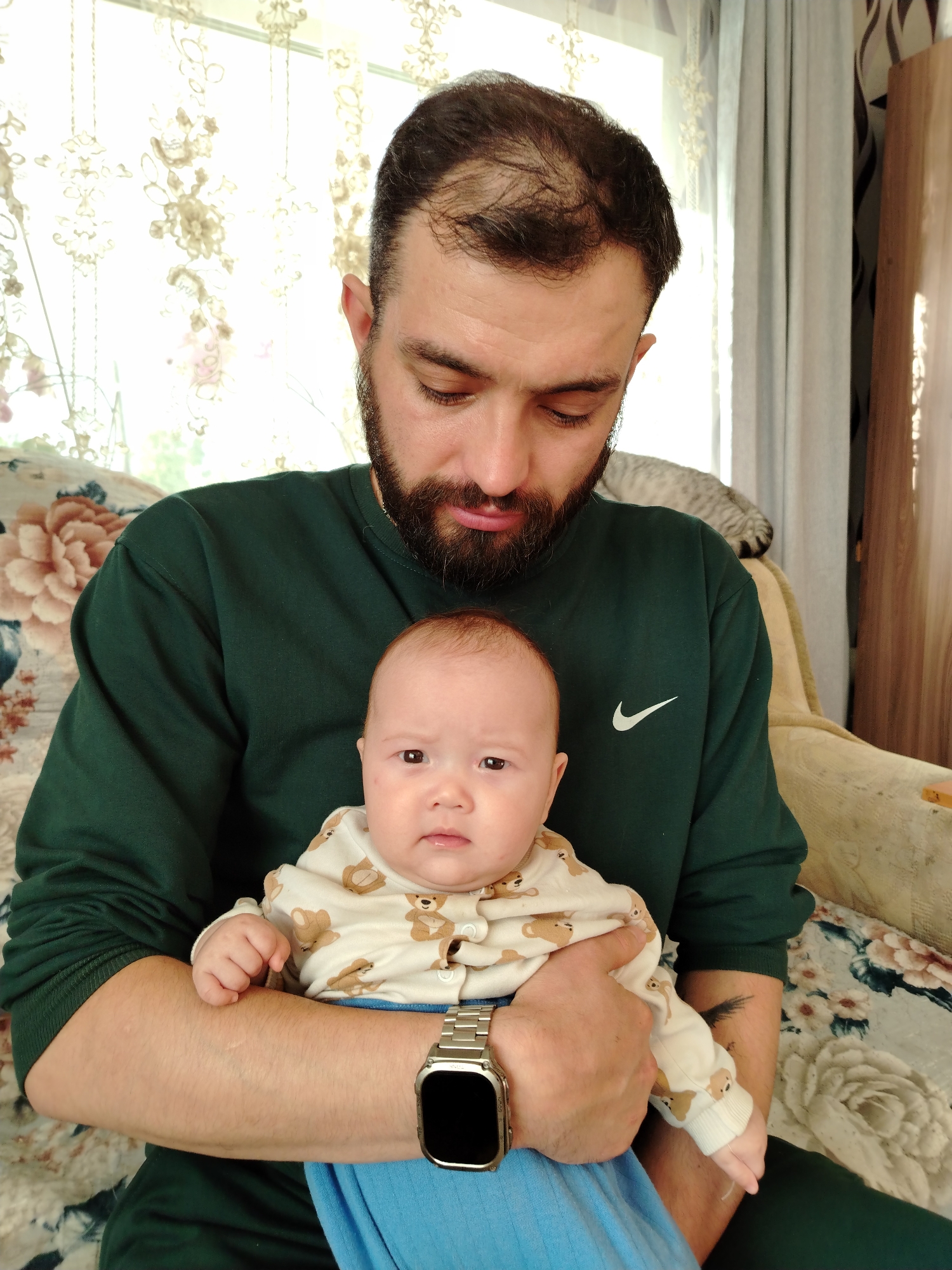 A man with a dark beard and receding hairline is holding a baby in his arms. The man is wearing a dark green sweatshirt with a white Nike logo on the upper right chest and a silver smartwatch on his left wrist. The baby is wearing a light-colored onesie with brown teddy bear patterns and blue pants. They are sitting indoors on a floral-patterned couch with a cat visible in the background on top of a tan chair. A patterned curtain is visible in the background. There are no readily apparent clues to indicate this is in the city of Гожан, Russia.