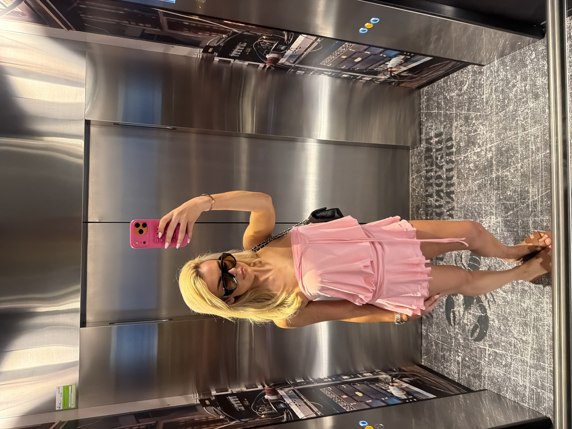 A blonde woman, wearing oversized dark sunglasses and a light pink strapless mini-dress, captures a mirror selfie inside an elevator. Her right arm is raised, holding a bright pink smartphone with an orange camera module. A black quilted shoulder bag with a chain strap hangs from her left shoulder, and a silver-colored bracelet is visible on her right wrist. Her fingernails are painted a light color. She wears flat, open-toed sandals. The elevator interior features reflective polished stainless steel walls. On the left and right sides, vertical panels display detailed black-and-white imagery of complex machinery or circuitry, with blue-lit circular buttons visible on the right panel. The dark gray floor is patterned with lighter gray abstract lines and features inverted text that reads "METROPOLITAN IRADIER". This text indicates the location is likely the Metropolitan Iradier fitness club in Barcelona, Spain.