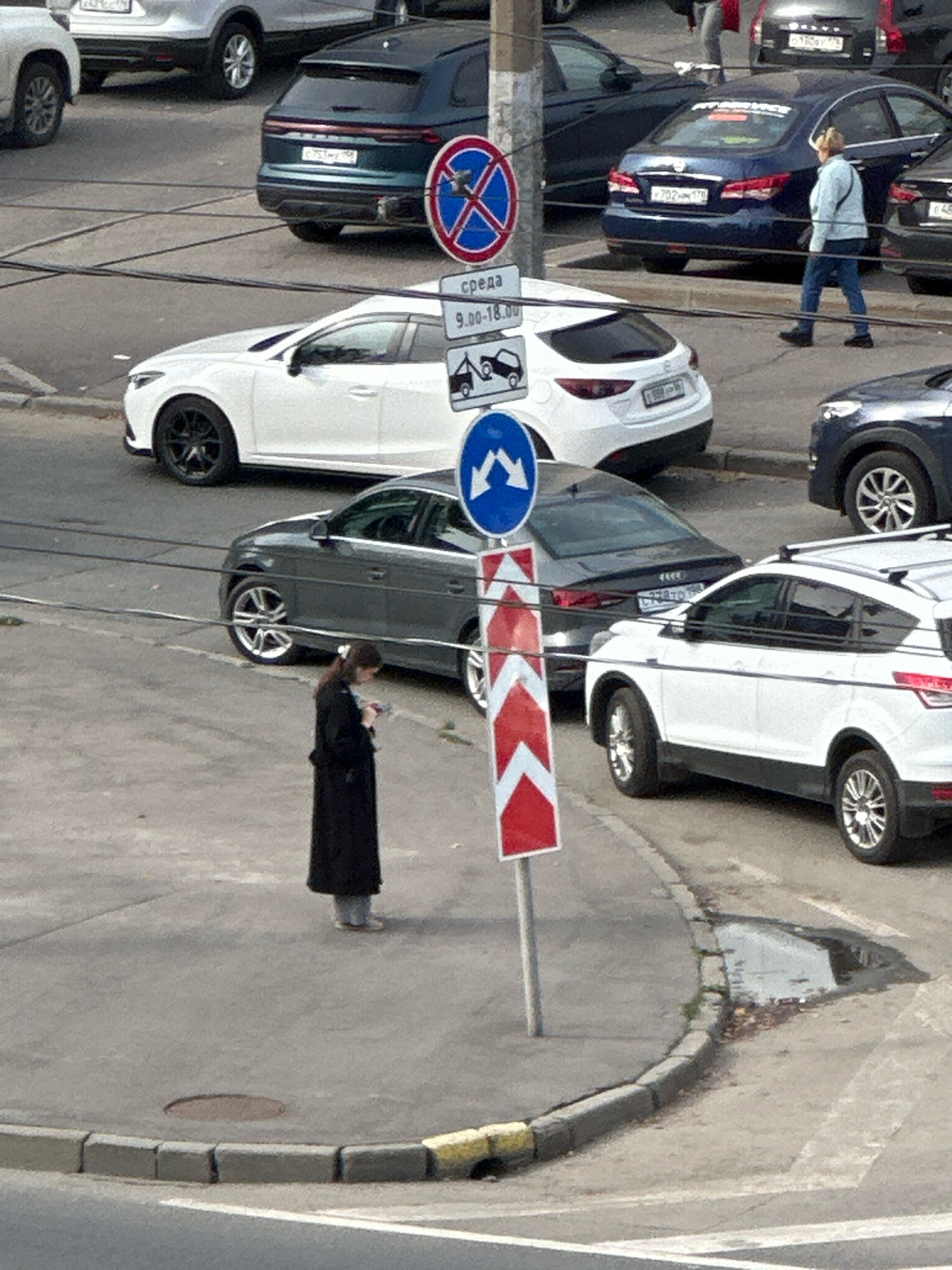 The image depicts a street scene in Saint Petersburg, Russia, with several cars parked and driving. In the foreground, a woman in a long black coat stands on a sidewalk, looking down at her phone. She is facing away from the camera. Behind her, a gray sedan is parked. In the midground, a white SUV is parked, and a dark blue sedan is driving away. Further back, a dark green SUV is parked, and several other vehicles are visible. Traffic signs indicating parking restrictions and directional information are present. The environment appears to be an urban street with paved roads and sidewalks, and some scattered debris on the ground. The lighting suggests it is daytime.