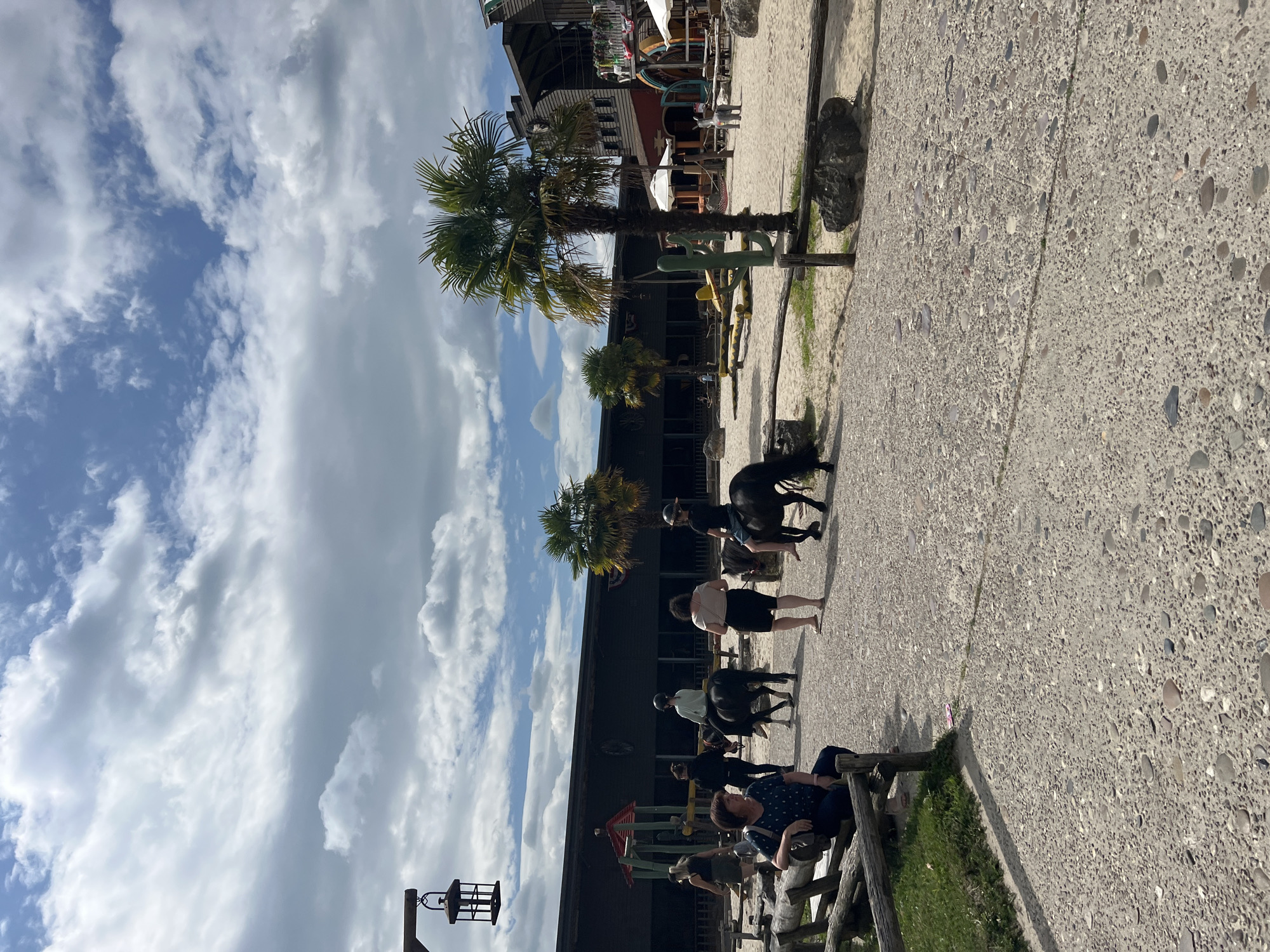 The image depicts an outdoor scene at a Western-themed park, likely a tourist attraction. In the foreground, a gravel pathway is visible, with a low wooden fence bordering a grassy area on the left. A woman is seated on a wooden bench in the grassy area, facing away from the camera. Several people are walking and riding ponies on the pathway and sandy area beyond.
In the middle ground, two ponies are being ridden by children wearing helmets. A group of adults is walking nearby. Behind them, there are more ponies, some appearing to be saddled and ready for riders. The environment includes sandy areas, wooden fences, and buildings that evoke a Western aesthetic, constructed with wood and dark siding. Palm trees and other vegetation are scattered throughout the area.
In the background, the park's structures are more prominent, featuring wooden facades, balconies, and signs. Some of these buildings appear to be shops or entertainment venues. The sky is partly cloudy, with a mix of blue sky and white clouds, suggesting fair weather.
The location is identified as the Western Village in Collendoorn, Netherlands. The scene suggests a lively atmosphere with families enjoying activities such as pony rides. The overall impression is that of a recreational area designed for entertainment and themed experiences.