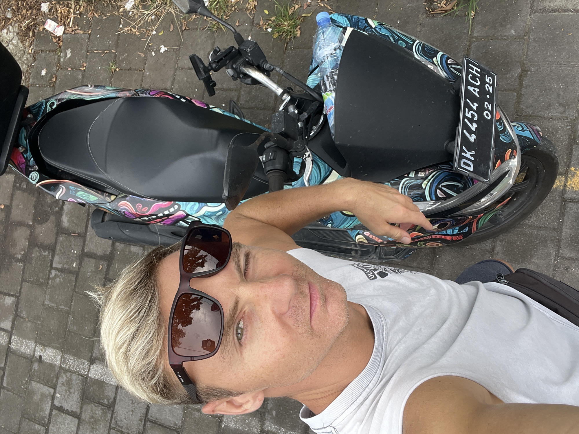 An adult male with blond hair, wearing dark brown Hugo Boss sunglasses, a white sleeveless shirt, and a dark fanny pack, takes a selfie while winking his left eye. He is leaning against a scooter customized with a colorful, abstract patterned wrap. The scooter features a black seat, handlebars with a phone mount, and a clear plastic water bottle in the front storage area. The scooter's black front license plate, visible at the bottom, reads "DK 4454 ACH" with an expiration date of "02-25". The scene is outdoors, on a grey interlocking brick pavement with scattered dried leaves and debris. The image is captured in the city of Celuk, Indonesia.