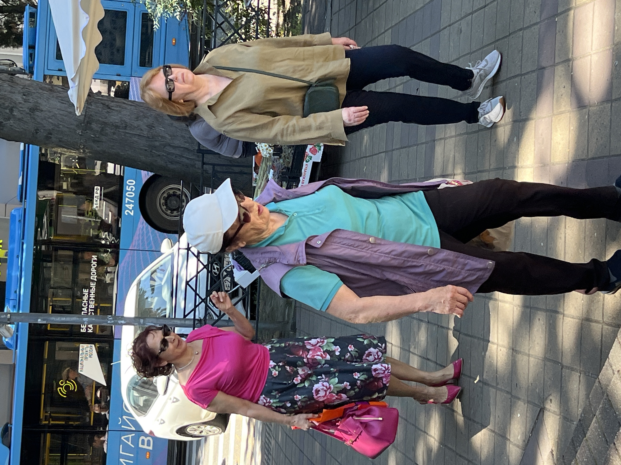 The image displays an urban street scene in Sochi, Russia, under bright daylight. Three women are walking from left to right on a patterned tiled sidewalk.
On the left, a woman with dark curly hair and dark sunglasses wears a fuchsia-pink short-sleeved top and a black midi-skirt adorned with large pink and red floral patterns. She wears matching fuchsia high-heeled shoes and carries a fuchsia handbag and an orange shopping bag in her left hand, with her right hand slightly raised.
In the central foreground, an older woman wears a white baseball cap, dark sunglasses, a teal short-sleeved polo shirt, an open purple vest, and dark pants. Her right hand is partially obscured, holding a light-colored object.
On the right, another older woman with reddish-brown hair and dark rectangular sunglasses wears an olive green long-sleeved collared shirt, dark trousers, and grey and white athletic shoes. A small dark green crossbody bag hangs from her right shoulder.
In the background, a light blue city bus is visible. Text on the bus windows reads "НАЦИОНАЛЬНЫЕ ПРОЕКТЫ РОССИИ" (National Projects of Russia) and "БЕЗОПАСНЫЕ КАЧЕСТВЕННЫЕ ДОРОГИ" (Safe Quality Roads). The bus number "247050" is painted near its rear wheel well. A white Hyundai sedan is parked or moving in front of the bus. A thick tree trunk stands prominently in the mid-ground, and a black metal fence extends across the scene. To the far right, a white market umbrella is partially visible, indicating an outdoor stall or cafe. The sidewalk is paved with square tiles, and distinct shadows suggest strong sunlight.