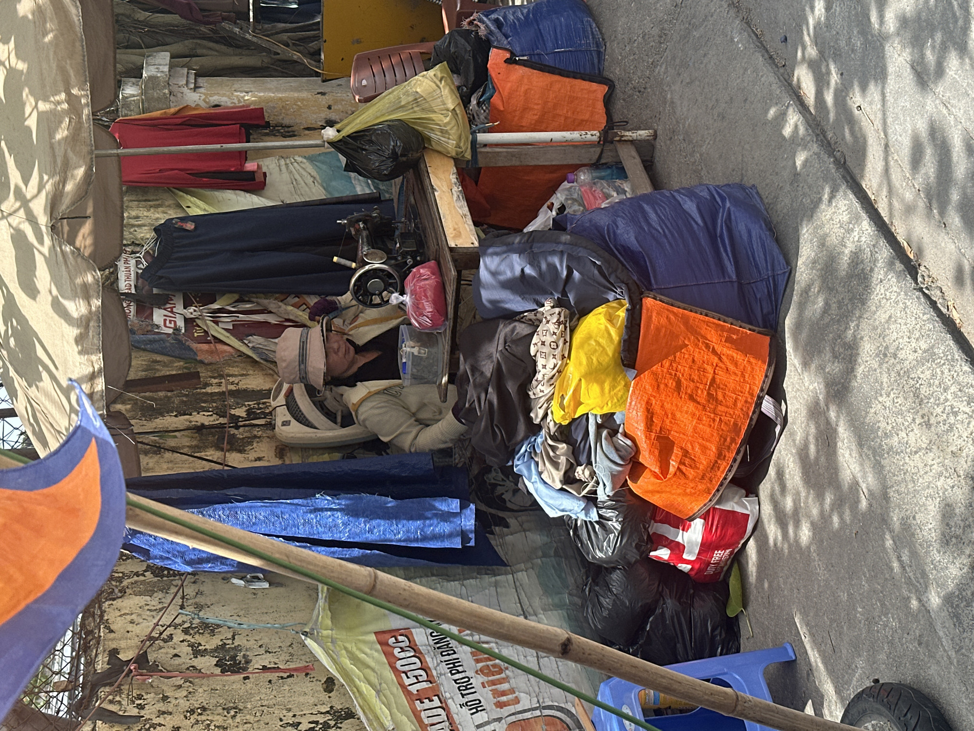 A person is sleeping outside next to a sewing machine in Bac Nha Trang Ward, Vietnam. The person is lying on a pile of belongings, including clothing, bags, and tarps. A red and white bag that reads "DUTY FREE" is visible. A banner that reads "ADE 150CC, HO TRO PHI DANG" is on the ground as well. There is a blue stool on the ground next to a wheel. Clothing hangs in the background. The area appears cluttered and makeshift.