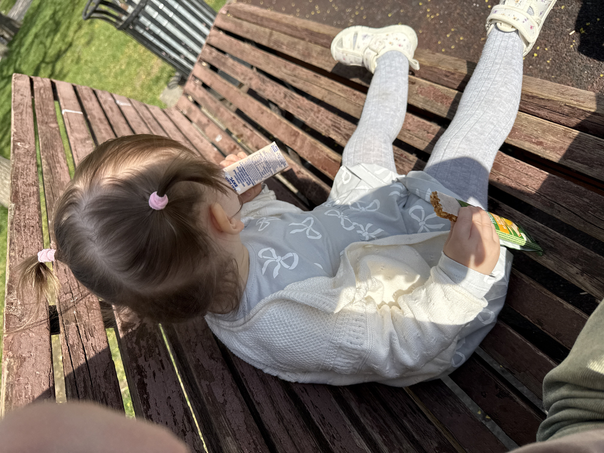 A young child sits on a wooden park bench in an outdoor setting. The child has light brown hair styled in two small ponytails with pink hair ties. The child is wearing light grey patterned leggings, a light grey dress with a white bow pattern, and a white knitted cardigan. On the feet are light-colored sandals with floral details.
The child is holding food items in both hands. One hand holds a squeezable pouch with nutritional information written in Russian. The other hand holds an open package of crackers or crisps.
The park bench has a slatted wooden design. In the background is a metal bench, green grass, and partial shadows of trees. The location is possibly in a park or recreational area within a city, but not definitively confirmed as Moscow, Russia. There are no clear identifiers confirming the city.