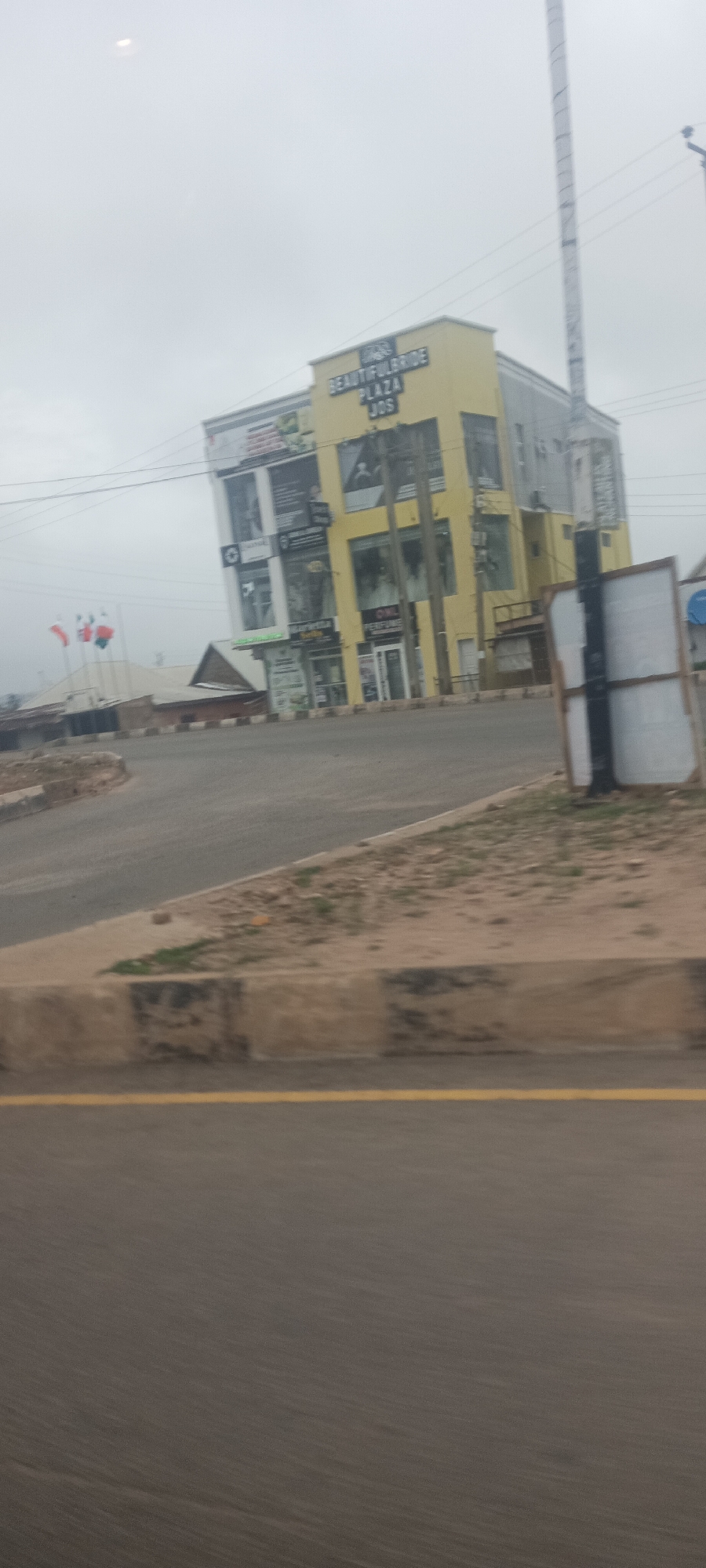 This eye-level, slightly angled photograph captures a street scene in Jos, Nigeria, featuring a prominent commercial building under an overcast sky. The multi-story building, which serves as the central subject, has a distinctive yellow upper section and a grey lower section on its main facade.
Visible text on the yellow part of the building clearly reads "BEAUTIFULBRIDE PLAZA JOS," indicating its name and location. The grey section of the building features multiple storefronts with various business signs, though most of the text on these signs is illegible due to resolution and distance, except for possibly "PERFUME" on one shop. A tall, thin pole, possibly a utility or antenna tower, stands to the right of the building, partially obscuring it.
In the foreground, a curved asphalt road with a faded yellow line is visible, bordered by a concrete curb and a dirt median with sparse vegetation. To the left of the building, in the distance, several flags on poles can be seen against the grey sky, with red and white colors being discernible. An obscured wooden signboard stands on the right side of the road, facing away from the viewer.
The overall lighting is flat and diffused, consistent with an overcast day, and there are no discernible shadows. No people or vehicles are visible, suggesting a quiet moment on the street. The scene depicts a typical commercial area in an urban setting.