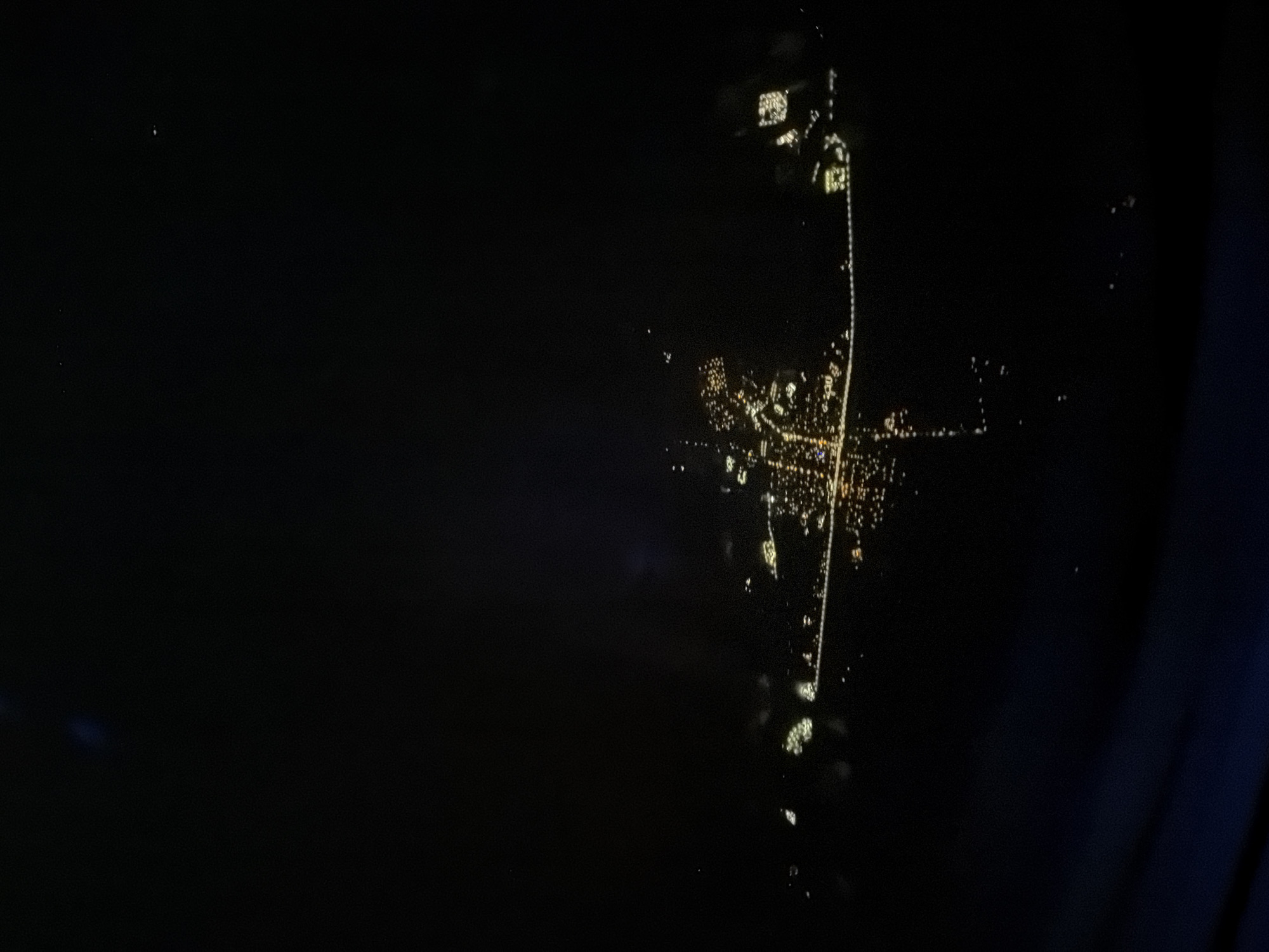 The image shows an aerial view of a city at night, likely taken from an airplane window. The city is recognizable by its streetlights and building lights, which form a distinct pattern against the dark background. A prominent north-south linear pattern of lights suggests a major thoroughfare or highway running through the city. The city appears to be Lordsburg, New Mexico, United States.
