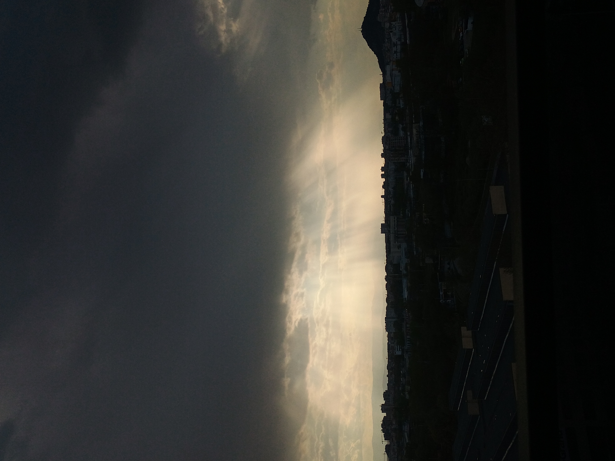 The image depicts a cityscape under a dramatic sky. To the left, dark storm clouds dominate, while to the right, shafts of sunlight break through lighter clouds, illuminating a densely built urban area. The location is identifiable as Plovdiv, Bulgaria, based on the presence of a distinctive hill in the background, consistent with the city's topography. Buildings of varying heights line the horizon, suggesting a mix of residential and commercial structures. The foreground shows a dark or shadowed area, possibly an indoor space, further framing the outdoor scene.