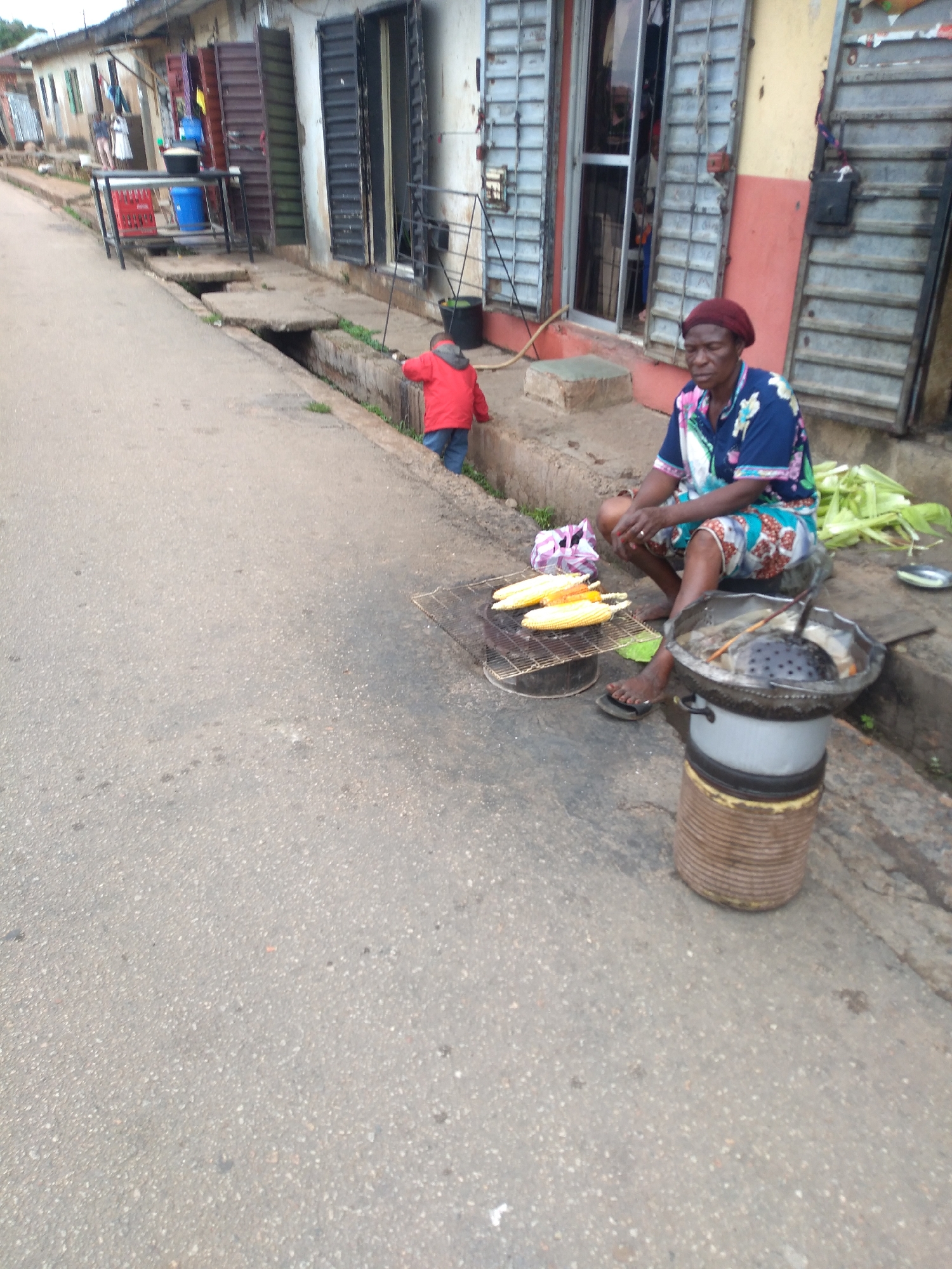 In Jos, Nigeria, a street vendor sits on the ground near her cooking setup. She is wearing a colorful patterned shirt and a red headscarf. In front of her, corn is being grilled on a metal rack over a fire. To her right, a pot with a slotted ladle sits on top of a makeshift stove. Behind her, a pile of corn husks is visible. A young child in a red jacket is standing in the street nearby, looking towards the shops lining the road. The shops appear to be typical of a commercial area in Jos, with metal shutters and awnings. The road is paved with asphalt and has a drainage ditch running alongside it. The overall scene depicts a typical street vendor operation in a bustling urban environment.