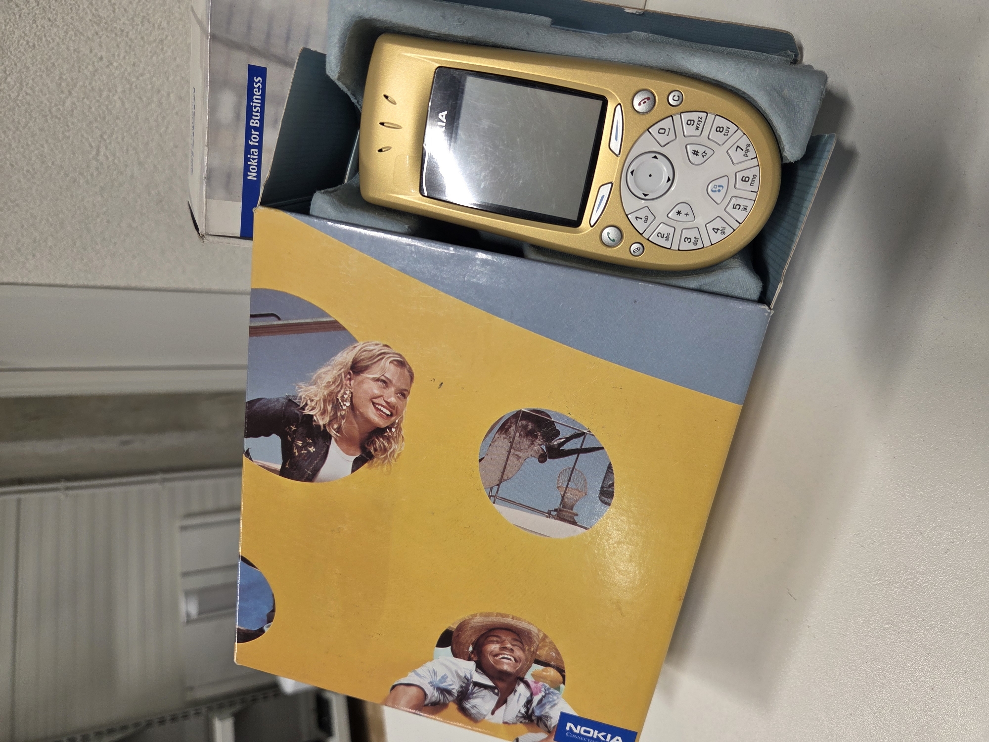 The image shows a gold Nokia 8800 Sirocco Edition mobile phone inside its retail packaging. The packaging is partially open, revealing the phone nestled in a blue foam insert. The box itself has a yellow lid with circular cutouts displaying images of people. The phone features a slider mechanism, a metallic finish, and a keypad with silver and grey buttons. A portion of a white box with the text "Nokia for Business" is visible in the background. The scene appears to be indoors, possibly in a retail or storage environment, with a white surface in the foreground. There is no discernible location within the city of Vilnius, Lithuania.