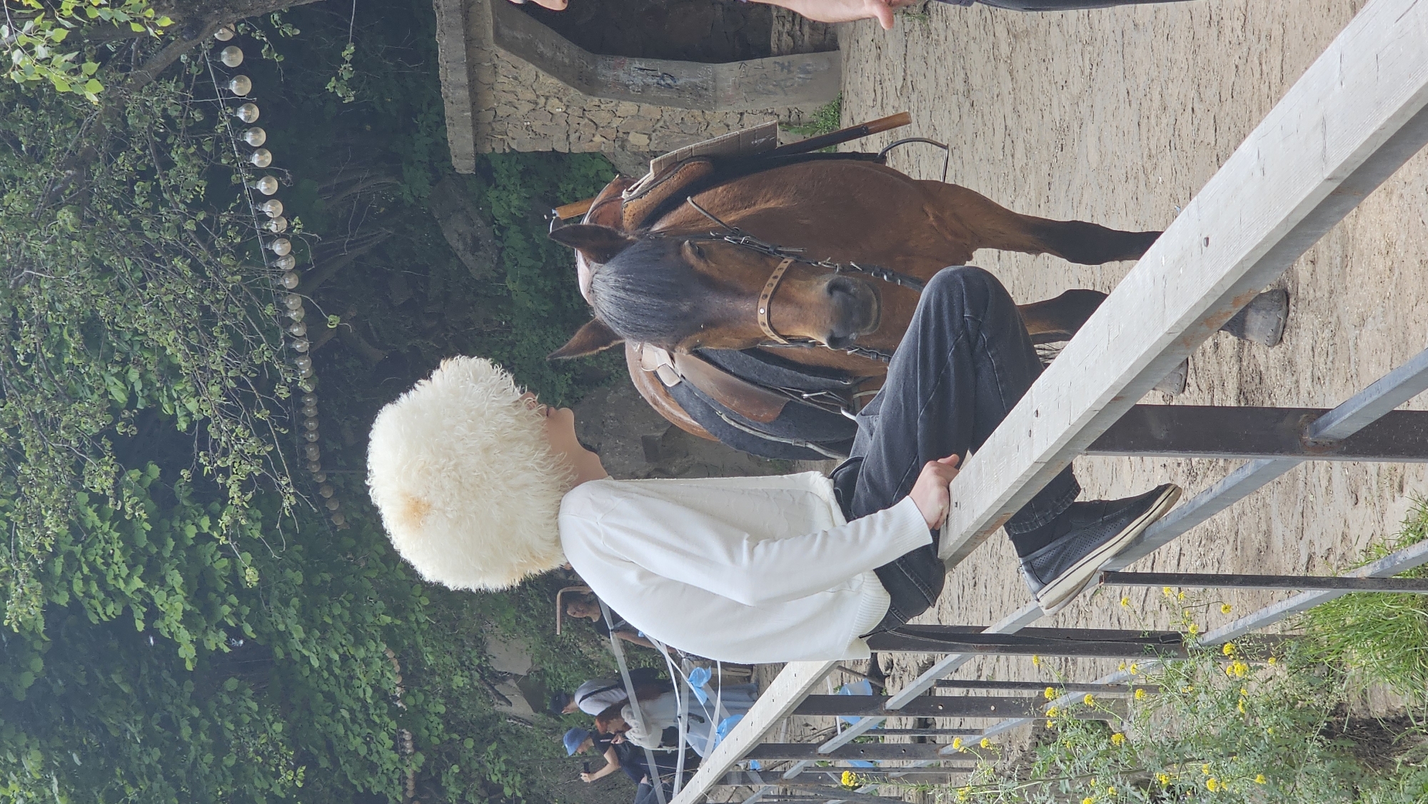 A young person wearing a fluffy white hat and a white long-sleeved shirt is perched on a wooden railing, facing away from the camera. They are wearing black jeans and dark shoes. Beside them stands a brown horse, saddled and bridled. The horse's head is turned towards the viewer, and its ears are perked. In the background, lush green trees and foliage create a natural setting. A string of lights is visible hanging from a tree branch. Other people can be seen in the distance, some taking photos. The ground appears to be dirt or sand. The overall scene suggests an outdoor recreational area, possibly a stable or park. The specific location within Красный Курган, Russia, is not determinable from the visual information.