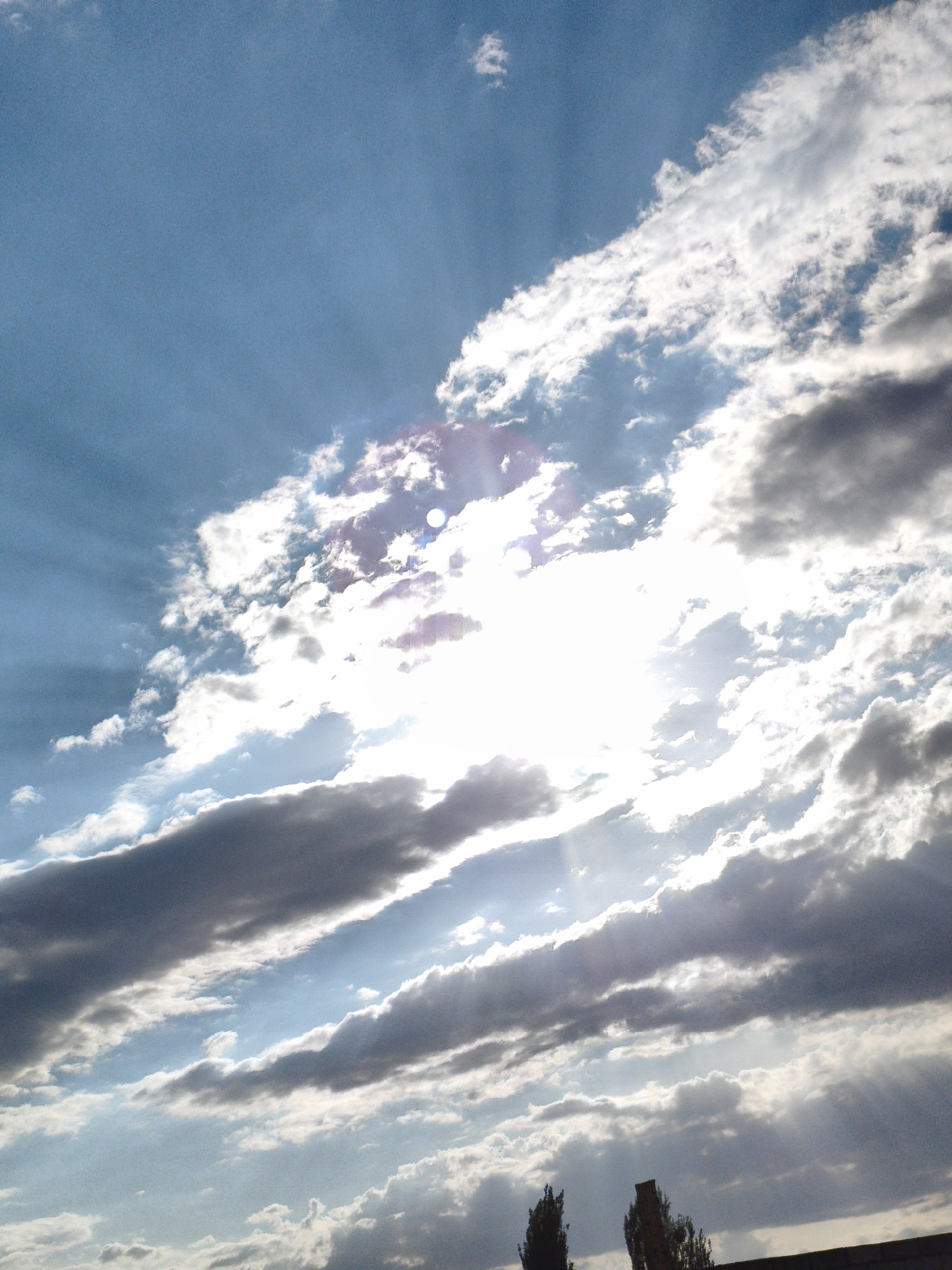 This is a low-angle, full outdoor shot of a sky with dramatic clouds and sunbeams. The sun is shining brightly through a gap in the clouds, creating visible rays of light that spread across the sky. The clouds vary in density and color, with some appearing bright white and others a darker gray. The overall impression is of a dynamic and possibly dramatic sky, as if during or after a storm. The bottom edge of the image shows the silhouette of a building or wall, and two trees. The location is in the city of Unknown, Unknown.
