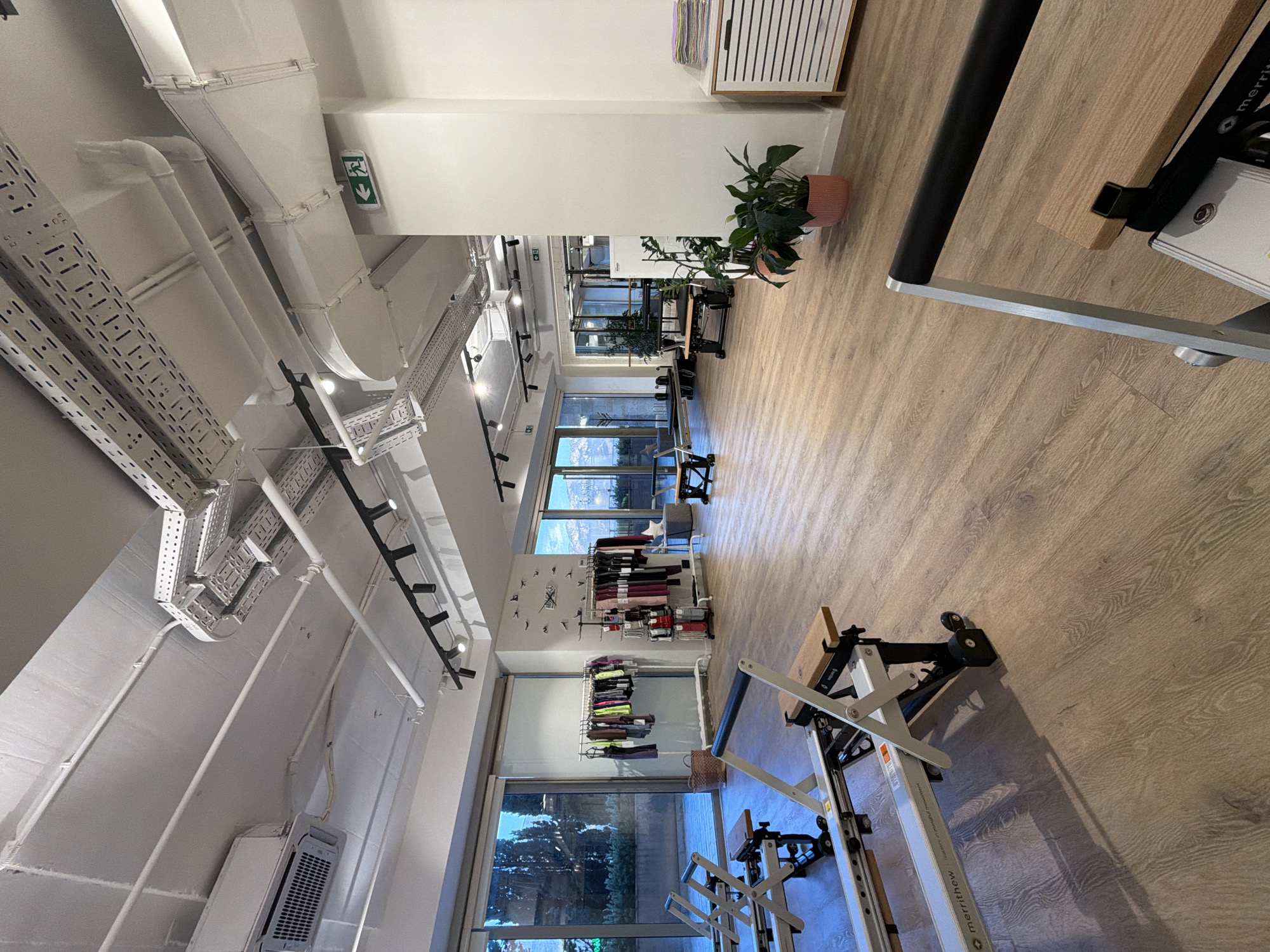 This is a medium-shot, interior photograph of a modern fitness studio, likely a Pilates studio, located in Boreti, Montenegro. The studio features a polished wooden floor and a high ceiling with visible industrial-style ventilation and lighting systems. In the foreground, two white Merrithew Pilates reformers are positioned, facing towards the left side of the frame. Each reformer has a wooden seat and metal frame with wheels.
The background reveals large windows offering a view of an outdoor landscape with trees and mountains, suggesting a scenic location. Racks of athletic apparel are displayed against a wall, with one rack featuring colorful leggings and tops, and another displaying darker toned activewear. Other exercise equipment, such as what appears to be a Pilates chair and reformer beds, are visible further in the background, along with potted plants. A white storage cabinet with ribbed doors is present on the right side of the image, topped with stacks of folded towels or mats. An illuminated green exit sign with a walking figure is affixed to a column on the right side of the ceiling. The overall ambiance is clean, bright, and organized, indicative of a professional fitness facility. There are no people present in the image.