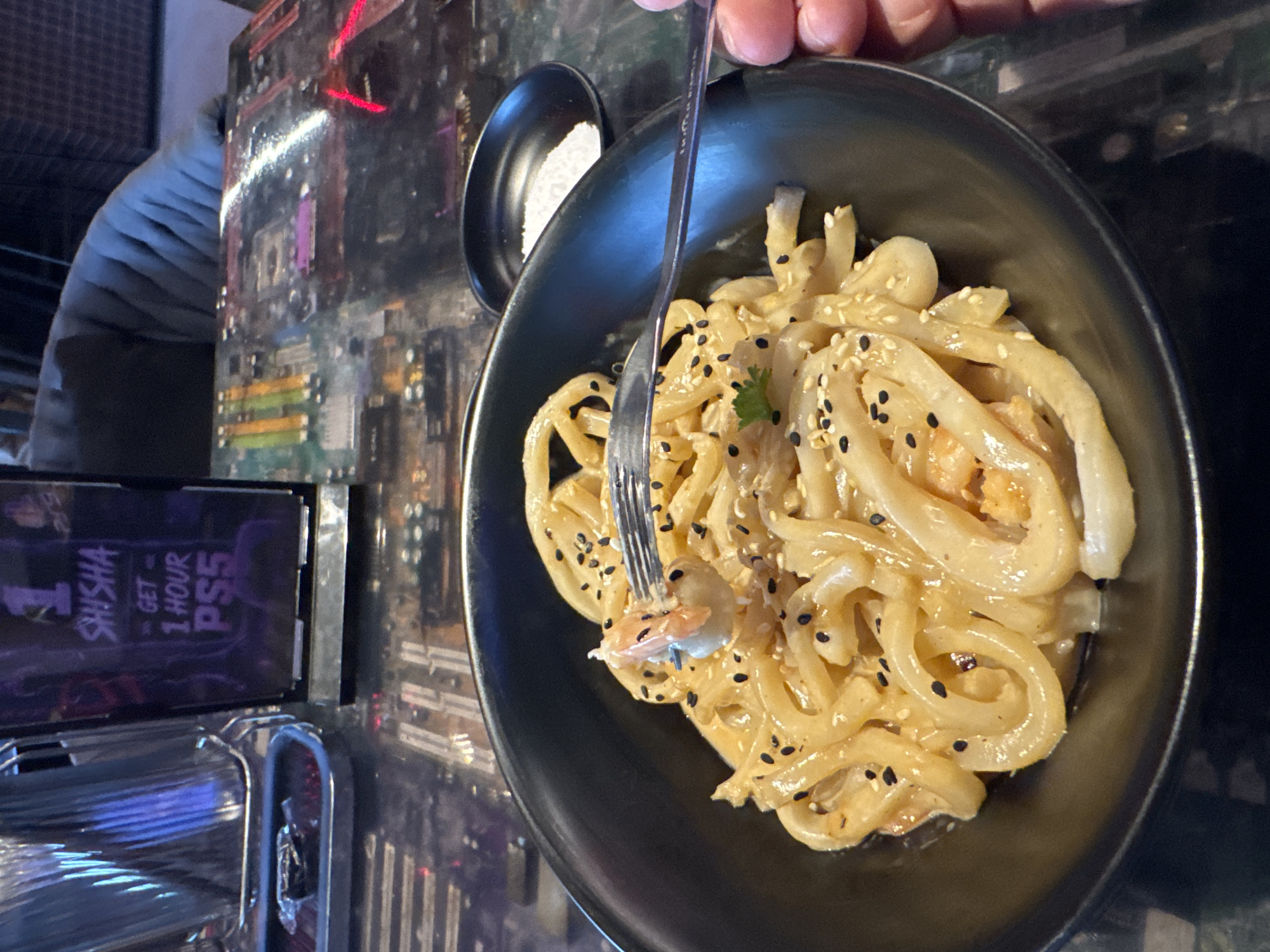 A close-up, eye-level shot shows a black bowl of udon noodles with shrimp and sesame seeds. A silver fork is lifting a piece of shrimp and noodle from the bowl. To the right of the bowl is a small black dish containing white seasoning. The table surface is dark and appears to be a mosaic of circuit boards. In the background, a vertical sign displays "1 SHISHA GET 1 HOUR PS5" in purple lettering. To the left of the sign, a metallic silver container with vertical ridges is visible. The overall lighting is dim.