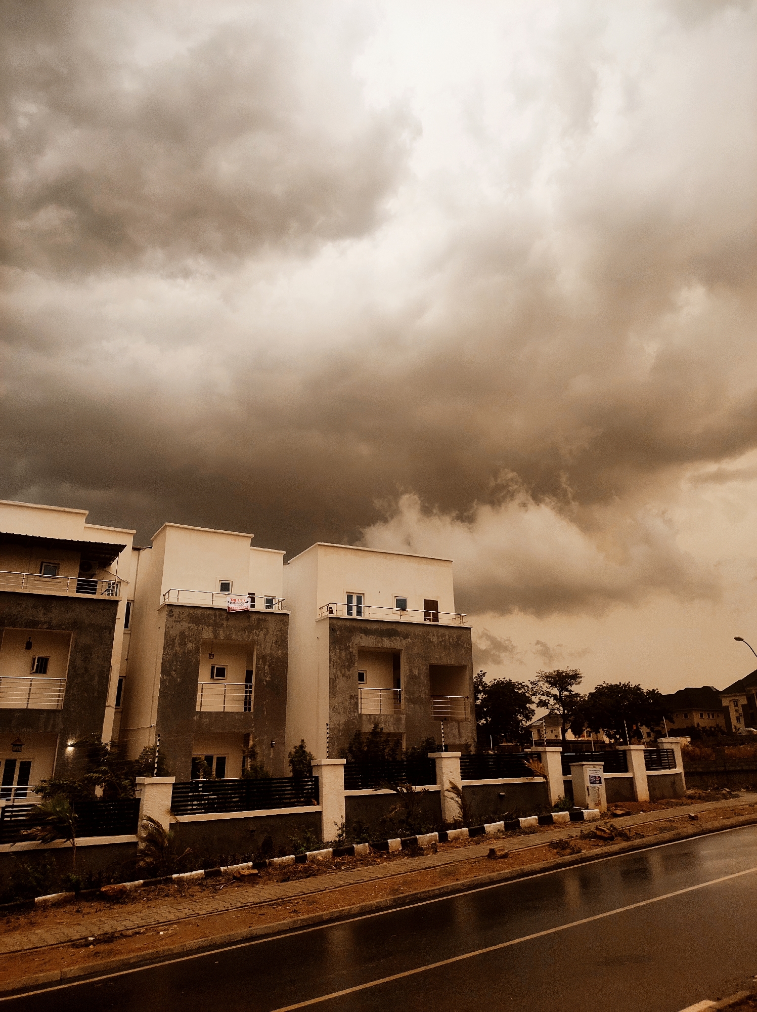 The image shows a street scene in Abuja, Nigeria, under a stormy sky. Three modern, two-story buildings are visible, likely residential units or townhouses. Each has a small balcony. A low fence runs along the front of the properties. A wet road is in the foreground, suggesting recent rain. There is a utility pole in the background, along with several houses. The overall tone of the image is sepia, lending it an aged appearance. There are no visible people.