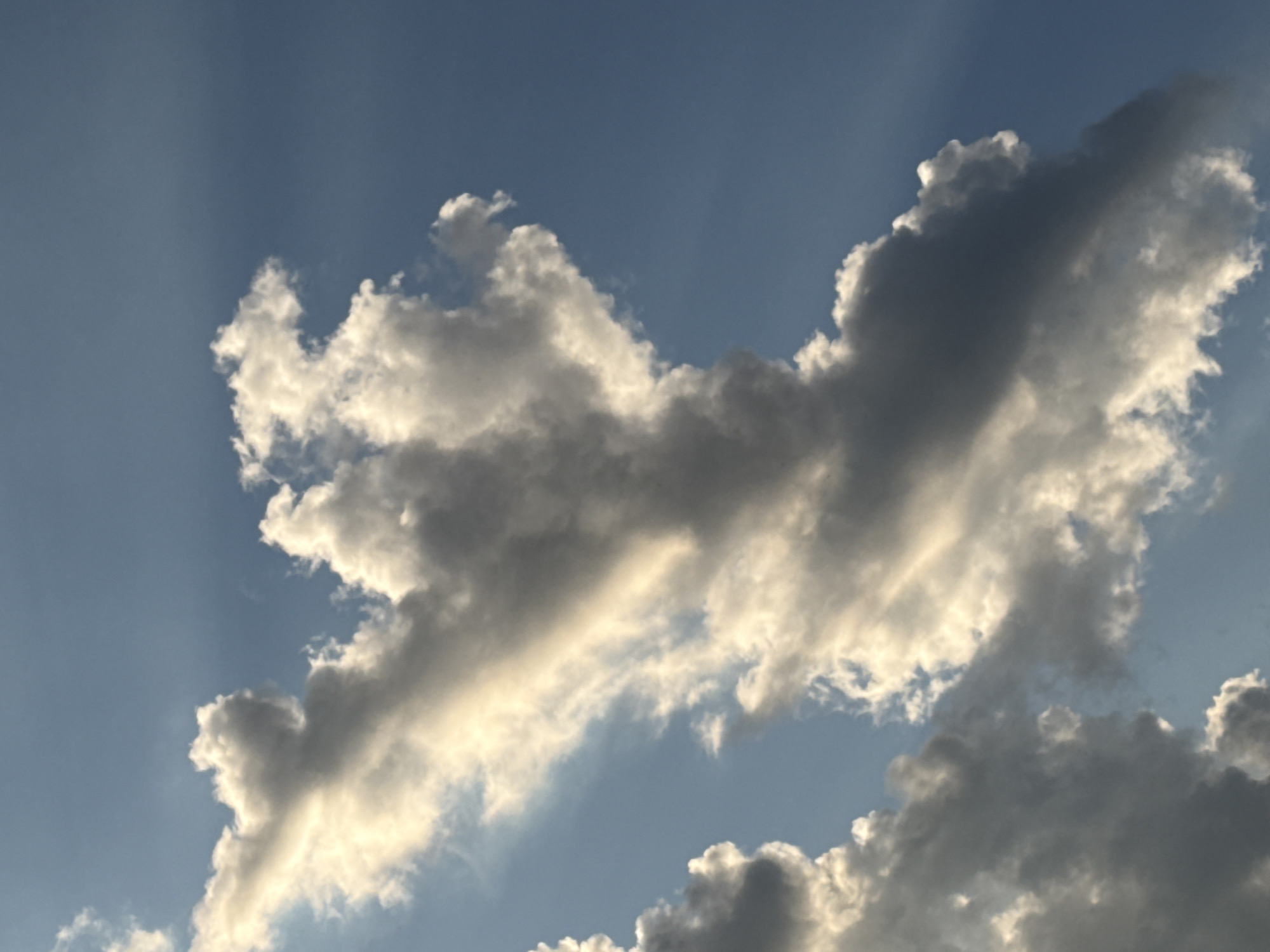 A vertical orientation photograph captures a dynamic sky over Unknown, Unknown. The scene is dominated by large cumulus cloud formations against a blue sky, illuminated by sunlight. A significant, elongated cloud mass, exhibiting both brightly sunlit edges (appearing white to pale yellow) and shadowed interior sections (appearing dark gray), extends diagonally across the frame from the lower-left to the upper-right. Visible crepuscular rays of sunlight penetrate the atmosphere, originating from behind the primary cloud mass, most clearly discernible in the upper-right quadrant and mid-left. The sky in the top-left corner is a lighter shade of blue, while the top-right shows a deeper blue with pronounced light rays. No human subjects, man-made objects, or ground features are present in the frame.