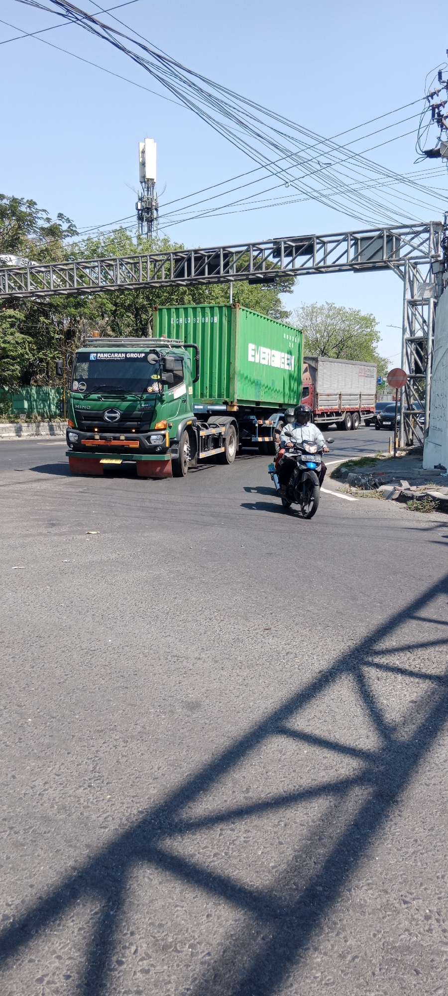 The image depicts a street scene in Gending, Indonesia, under a clear blue sky. A large green shipping container is being transported by a truck bearing the "PANCARAN GROUP" logo and "HINO" branding on its cabin. The container itself is labeled "EVERGREEN." To the right of the truck, a motorcycle carrying two individuals is in motion. In the background, a cell tower is visible, along with overhead power lines and a metal overhead structure, possibly part of a bridge or overpass. The road surface is asphalt, with some debris scattered on it. Shadows of the overhead structure are cast on the road. Another truck and a black car are also present further down the road.