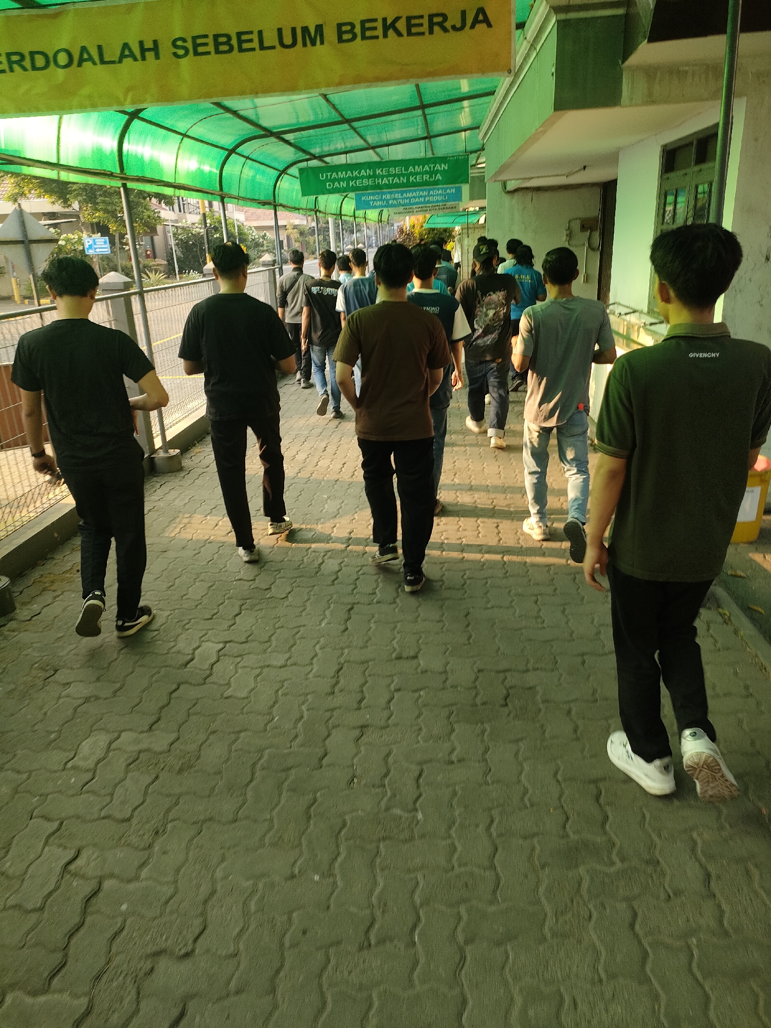 The image depicts a group of young men walking along a paved pathway under a green, covered walkway. The walkway is illuminated by natural light, casting long shadows on the ground. A yellow banner with Indonesian text, which translates to "Pray Before Working," is visible overhead. Further along, a green sign with white text announces "Prioritize Safety and Health" and other safety instructions. The men, dressed in casual attire such as t-shirts and pants, are walking in single file away from the camera. The environment appears to be an outdoor area, possibly a campus or industrial facility, given the presence of buildings and fences in the background. The paving stones on the walkway are a distinctive interlocking pattern. The overall scene suggests a group of individuals heading to or from a work or educational setting.