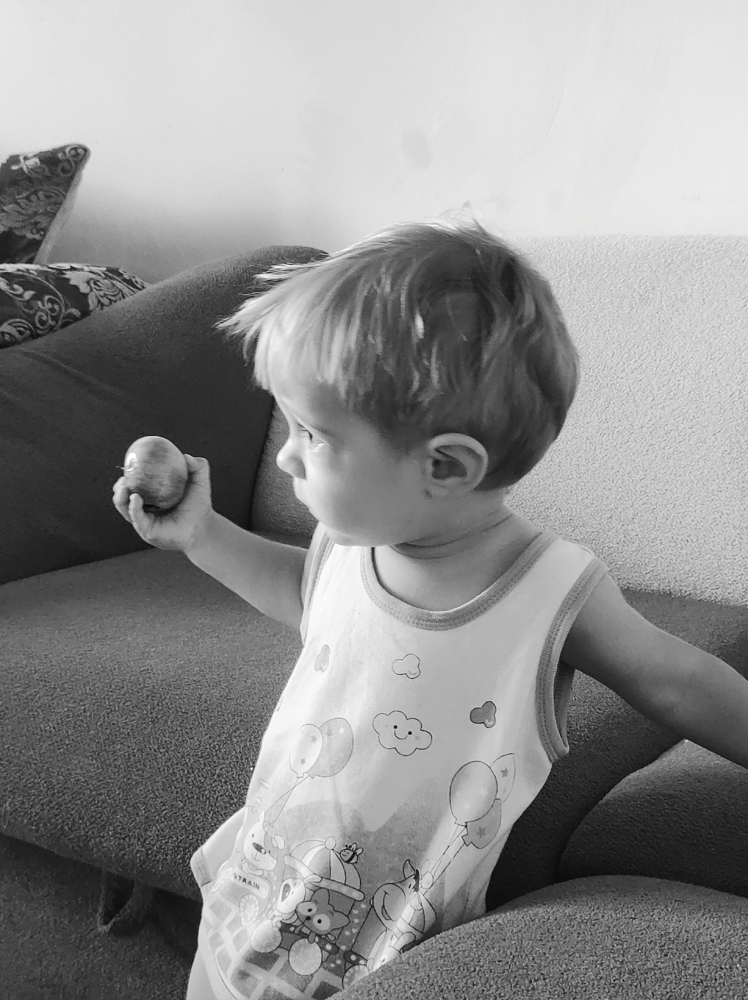 In a black and white, medium-shot, the upper body of a young Caucasian child is visible, seated on a couch. The child is looking to the right with their head turned away from the camera, holding a small round object, possibly an apple, in their right hand. The child is wearing a sleeveless shirt with a graphic print of clouds, balloons, and cartoon characters. The background consists of a couch with patterned pillows and a textured wall. The scene appears to be indoors, within a residential setting, potentially in the city of Fokino, Russia, given the provided context.