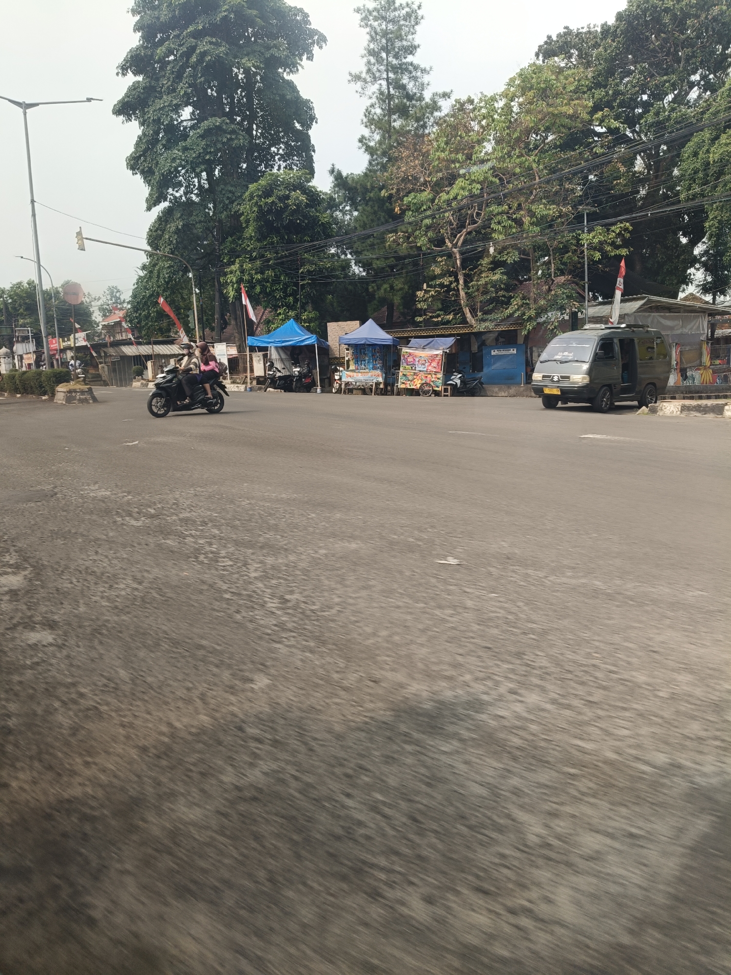The image depicts a street scene in Pandeglang, Indonesia, likely in an urban or semi-urban area, with a focus on the road and the surrounding environment. In the foreground, a road with visible texture and some wear is captured, suggesting it is a functional thoroughfare. A black motorcycle with two occupants is in motion on the road, with the rider wearing a pink shirt and the passenger wearing a dark top and a helmet. To the left of the motorcycle, a street light pole is visible.
Behind the road, a row of small stalls or kiosks are set up. These stalls appear to be temporary structures, some with blue tarpaulin roofs. One stall has colorful displays, possibly indicating merchandise for sale. A dark grey van is parked or slowly moving past these stalls. The van has tinted windows and a license plate. In the background, a large, mature tree with dense foliage dominates the upper left portion of the image, with other trees and vegetation visible throughout the scene. The sky is overcast, suggesting a cloudy day. The overall impression is a snapshot of daily life and commerce in a street setting.