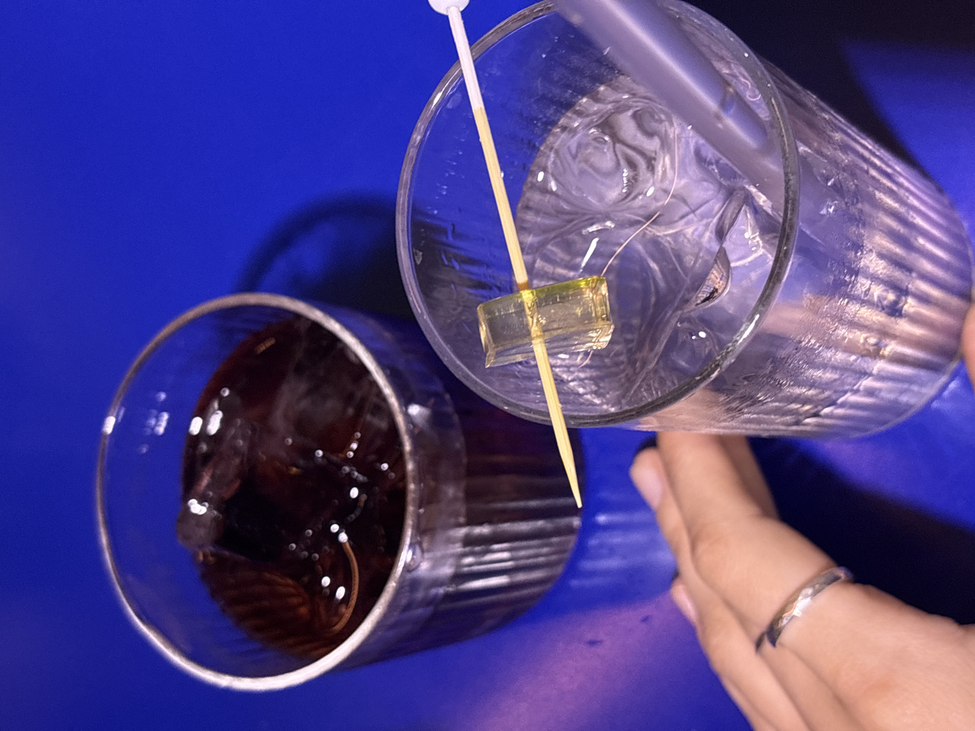 This is a close-up, overhead shot of two drinks on a vibrant blue surface, possibly a table. To the left, a glass filled with a dark liquid and ice is visible. To the right, another glass contains a clear liquid with ice and a straw. A wooden skewer with a clear, yellow cube-shaped garnish rests across the rim of the second glass, with the garnish submerged in the drink. A hand, adorned with a silver ring, is visible in the foreground, reaching towards the glass on the right. The lighting suggests an indoor setting, likely a bar or restaurant. There is no indication of the location being Perm, Russia.