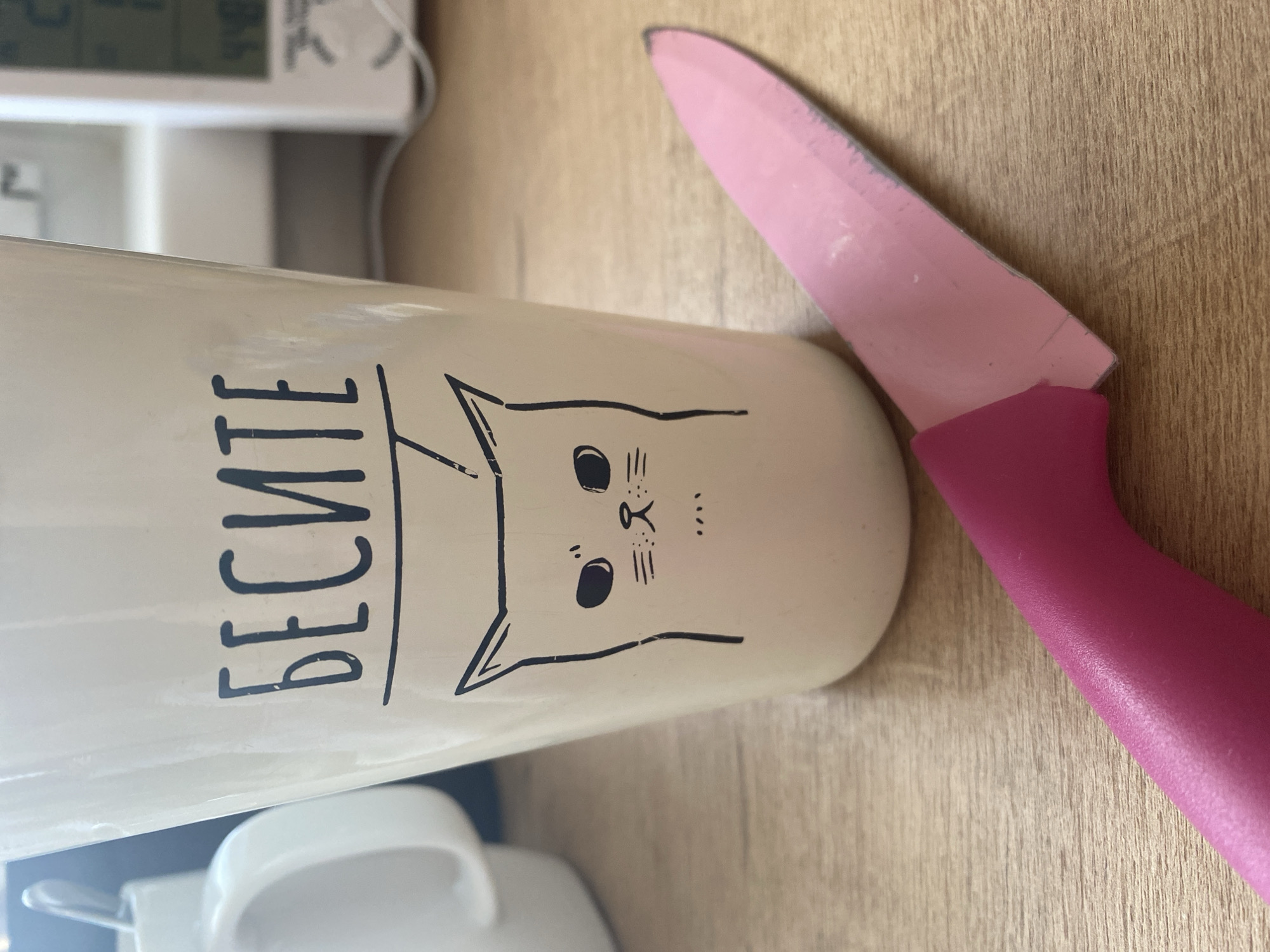 This image depicts a close-up, static scene set on a light-colored wooden surface, likely a countertop or desk, in Chaikovsky, Russia. The primary subjects are a tall, light-colored ceramic mug and a pink-bladed knife. The mug features a black line drawing of a cat's face with solid black eyes and small, horizontal lines for its nose and mouth. Above the cat, the Cyrillic word "БЕСИТЕ" (transliterated as "BESITE," meaning "you annoy") is printed in black uppercase letters with an underline. The knife has a vibrant pink handle and a matching pink blade, which shows visible wear, chipping, and areas where a darker material underneath the pink coating is exposed, particularly along the edge and tip. The knife is positioned with its handle in the lower-left foreground, extending diagonally towards the upper right. In the blurred background to the left, part of a white ceramic mug or cup with a handle is visible. To the right in the blurred background, a white digital device, possibly a thermometer or clock, displays numbers (e.g., "448") and symbols. No individuals are present, and no active interactions are occurring.