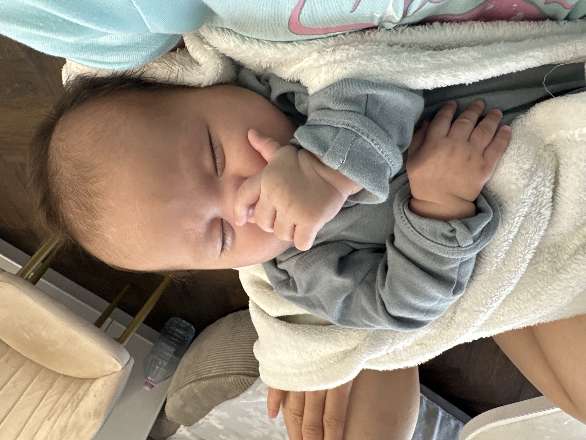 A baby is asleep in an adult's lap. The baby is wrapped in a white blanket and wearing a gray, long-sleeved onesie. The baby's face is visible, and one hand is near its mouth. The adult is wearing a light blue sweater. The background shows a beige armchair, a water bottle, and flooring. Location: Unknown, Unknown.