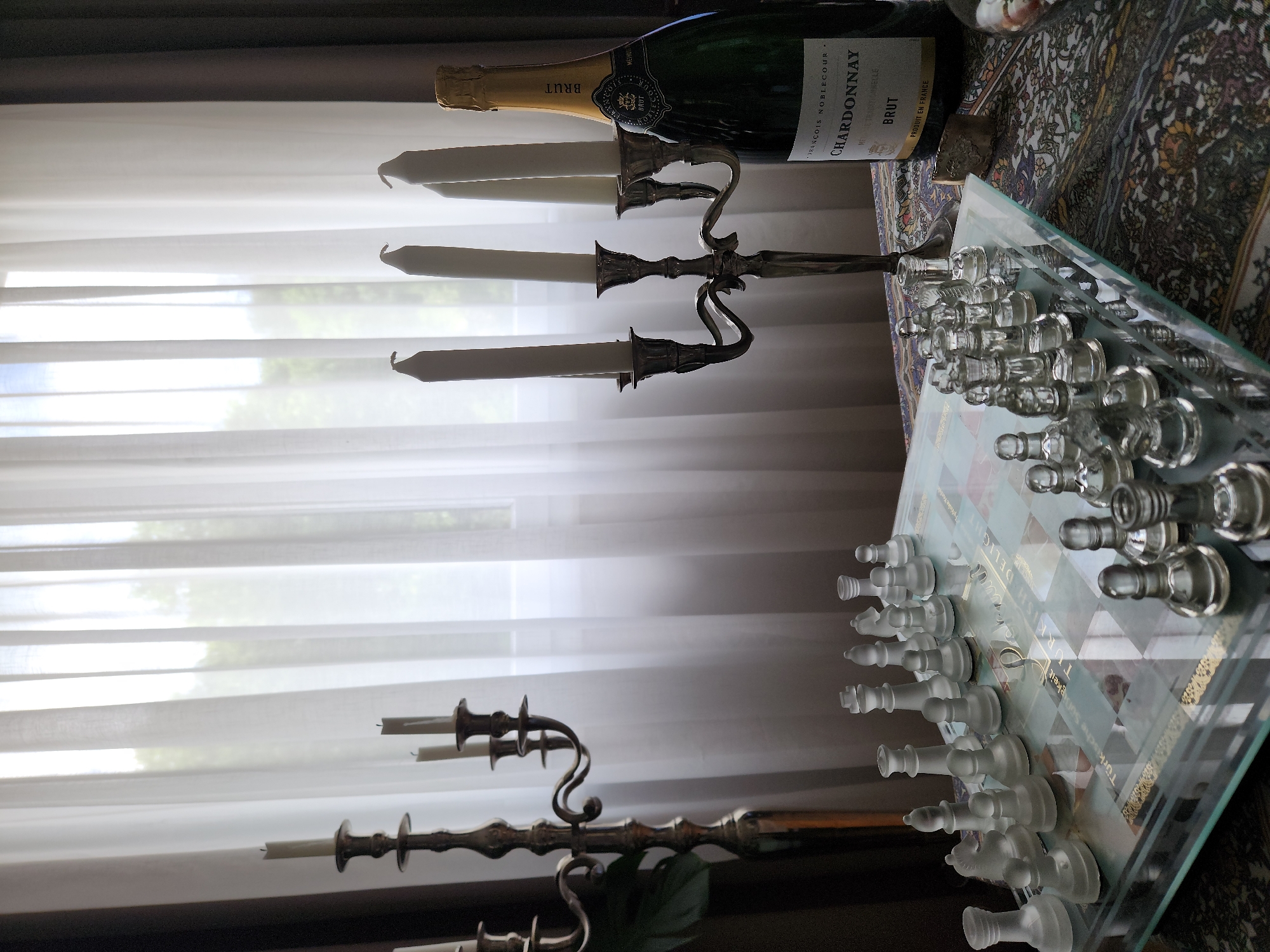 This image depicts an indoor scene located in Idar-Oberstein, Germany. In the foreground, a rectangular glass chessboard is visible, populated with two full sets of translucent glass chess pieces, one frosted and the other clear, arranged in their starting positions. The center of the chessboard features decorative text, including "Turkish Delight" and "Lokum."
To the left, a tall, ornate bronze-toned candelabra with three arms holds three unlit white taper candles. To the right, a shorter, four-armed silver-toned candelabra also holds four unlit white taper candles. Adjacent to the right candelabra stands a dark green champagne bottle, labeled "FRANÇOIS NOBLECOUR CHARDONNAY BRUT," with a gold foil cap. These items are situated on a table covered by a richly patterned fabric with intricate designs in various colors including blue, red, and gold.
The background is dominated by a large window covered with sheer, white translucent curtains, through which diffused natural light enters the room. Vague outlines of green foliage and a bright sky are discernible outside the window. No individuals are present in the image.