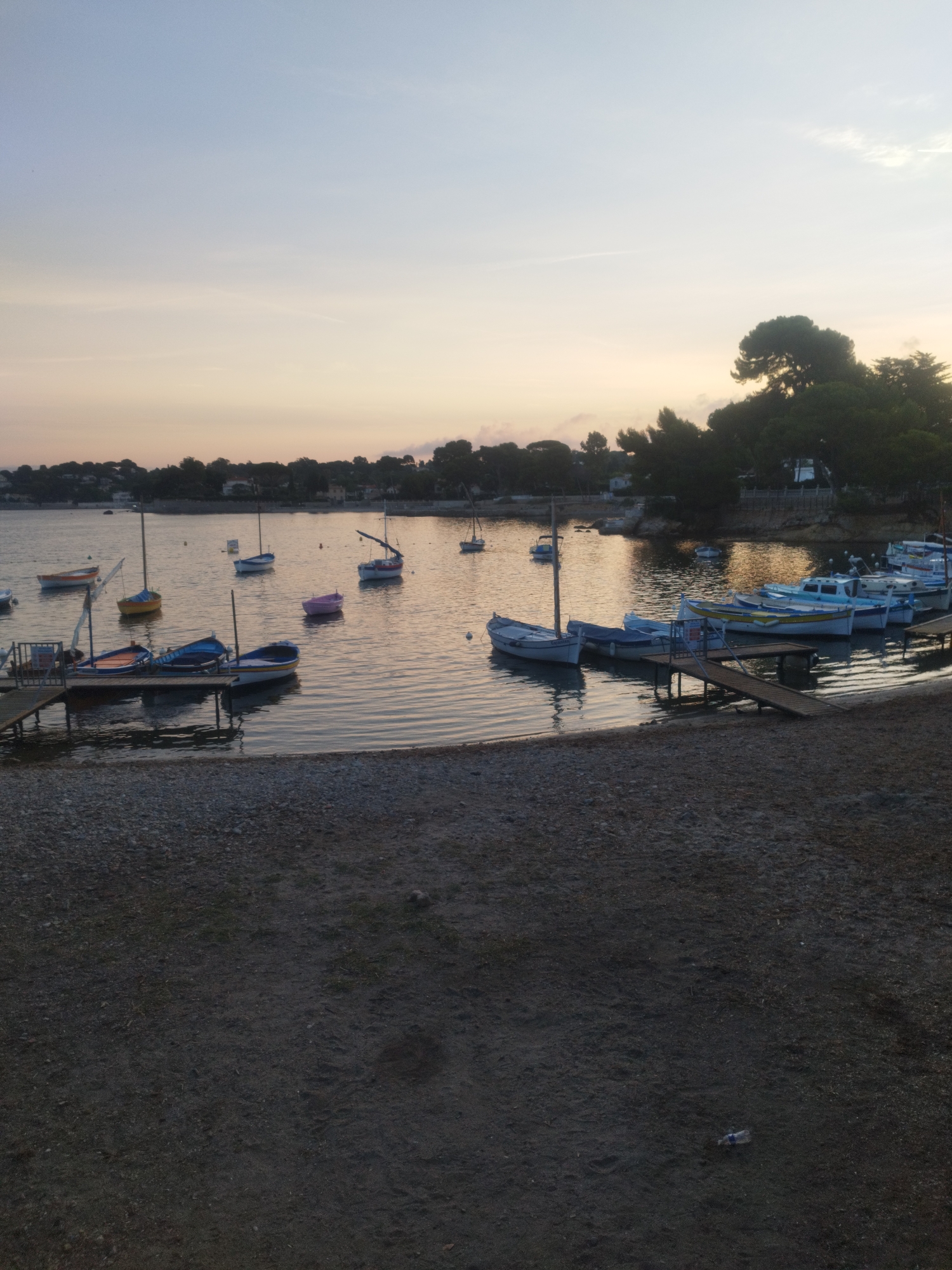 An offshore scene in Antibes, France, depicts a tranquil body of water dotted with several small boats. The sky above is a soft gradient of pale blue and warm orange, suggesting either dawn or dusk. Along the shoreline in the distance, a line of trees and buildings forms a gentle curve. In the foreground, a pebbly beach leads to the water's edge. The water is calm, reflecting the sky and the boats, creating a serene atmosphere.