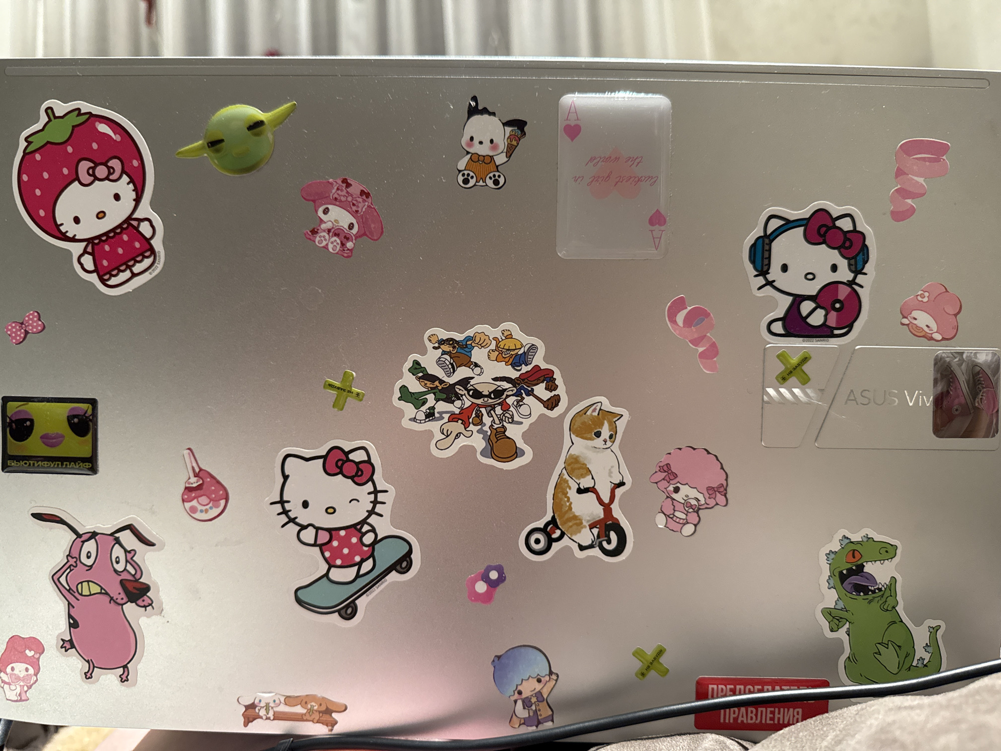 The image displays a collection of colorful stickers adhered to a silver laptop lid. The stickers feature various cartoon characters, including Hello Kitty in multiple designs, My Melody, Pochacco holding an ice cream cone, a cat on a tricycle, a pink poodle-like character, Courage the Cowardly Dog, a green dinosaur from Rugrats, and a group of characters from various anime. There are also abstract shapes like pink swirls and a playing card design. Text in Russian is visible on some stickers, including "БЬЮТИФУЛ ЛАЙФ" and "ПРЕДОСТЕРЕЖЕНИЯ". The laptop is placed on a soft, possibly fabric surface, and a portion of a black cable is visible at the bottom. The background appears to be an indoor setting with vertical blinds. The image does not provide any information about the city of Бараўлянскі сельскі Савет, Belarus.