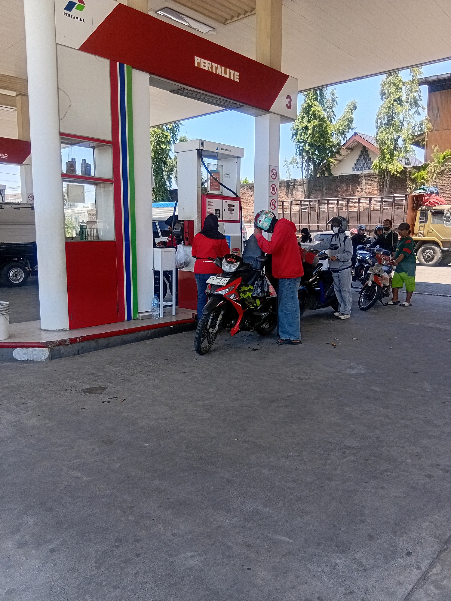 At a Pertamina gas station in Bendotretek, Indonesia, several individuals are present. A woman in a red jacket and blue jeans stands near a motorcycle, holding a plastic bag. A person wearing a gray hooded sweatshirt and light gray pants is attending to a black motorcycle. Another person, wearing a black helmet and gray clothing, is also near a motorcycle. In the background, several other individuals and motorcycles are visible, as well as a truck and a building. The scene is illuminated by daylight.