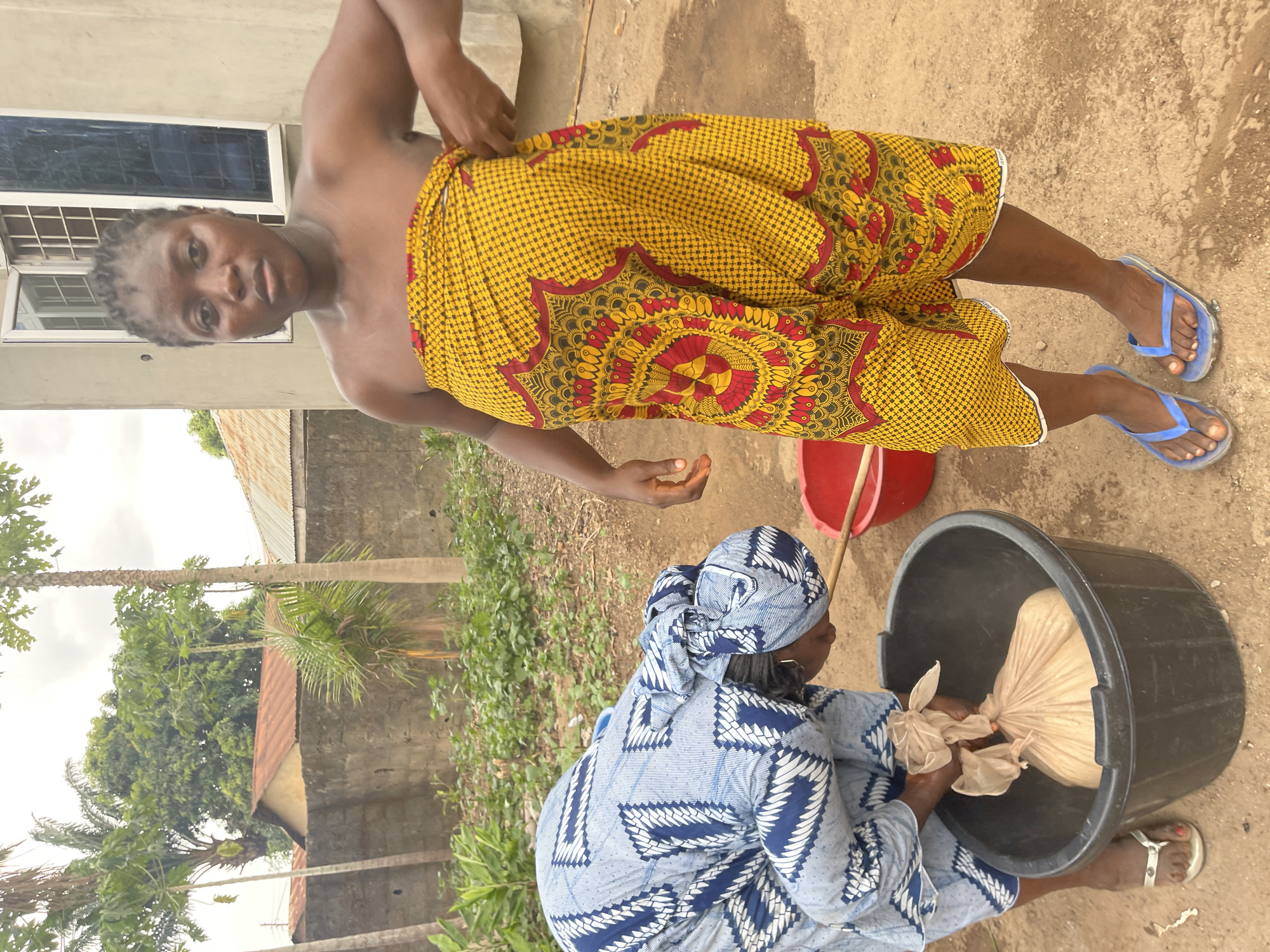 Here is a detailed description of the image:
**People:** Two dark-skinned women are the main subjects. One woman, standing, wears a yellow and red patterned cloth wrapped around her torso. She is barefoot and has her hair braided. The other woman, seated, wears a blue and white patterned garment and matching headwrap.
**Subjects:** The subjects are engaged in some kind of washing or cleaning process. The standing woman is looking at the camera.
**Objects:** A large black plastic tub filled with water contains a large bag. A red bucket sits nearby. The standing woman is barefoot wearing blue flip flops.
**Actions:** The seated woman is interacting with the bag inside the tub, possibly washing or cleaning it. The standing woman appears to be adjusting her clothing.
**Interactions:** The two women are in close proximity and appear to be working together on a task.
**Environment:** The scene takes place outdoors in a dusty environment. A building is visible in the background. Other buildings with rusty roofs and trees are also in view.
**Location:** While Akwanga, Nigeria is mentioned, there are no specific landmarks or distinct features that definitively identify the location within the city.