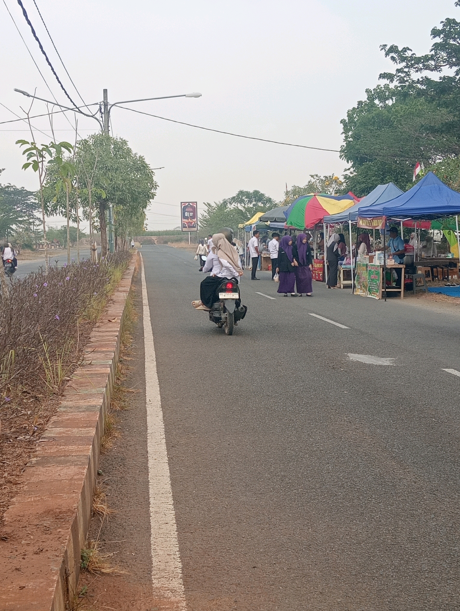 In Sidoharjo, Indonesia, a road is lined with market stalls under colorful tents. People, many wearing headscarves, are seen walking and sitting at the stalls. A person rides a scooter away from the camera. On the left side of the road, a grassy area with some plants is bordered by a low brick wall. Overhead, power lines cross the sky, which is overcast. A street lamp is visible, and a billboard with the number "81" is in the distance.