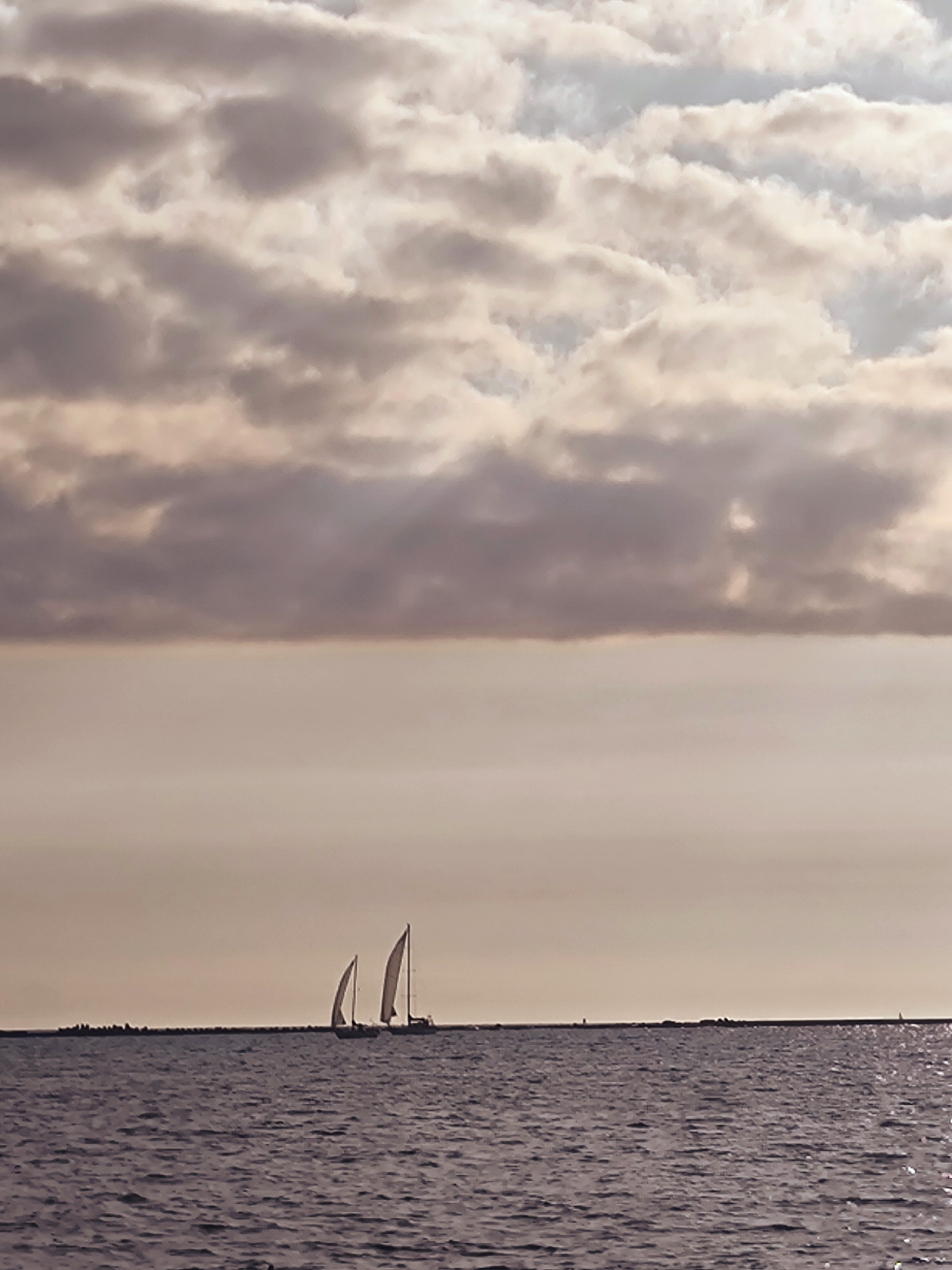 The image depicts a tranquil sea scene with two sailboats navigating the water. The sky is filled with dramatic, layered clouds, tinted with shades of pink and gray, suggesting either dawn or dusk. The water is a deep, calm blue, with gentle ripples reflecting the muted light. A long, low-lying landmass or breakwater stretches across the horizon, dotted with faint structures that could be buildings or navigational markers. The overall atmosphere is serene and vast, with the sailboats appearing as small, elegant figures against the expansive backdrop of sea and sky. The scene is likely a coastal area, possibly near a harbor or bay, offering a sense of open space and natural beauty.
