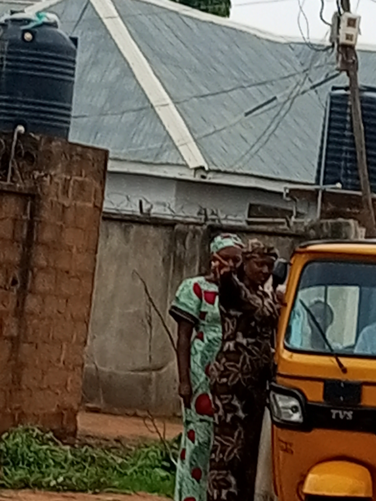Two women stand near a yellow auto-rickshaw in Tempure, Nigeria. The woman on the left wears a light green and white patterned dress with red accents, and a matching headscarf. The woman on the right wears a dark brown and white patterned dress and a headscarf. The auto-rickshaw has "TVS" branding on its front. In the background, a large black water tank sits atop a brick structure, and a corrugated metal roof extends across the scene. Power lines and a utility pole are visible above the roof. A grey concrete wall with barbed wire runs horizontally across the midground. Green vegetation grows at the base of the brick structure.