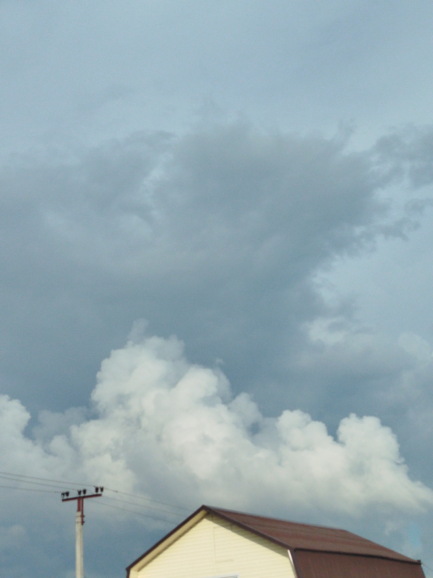 The image depicts a cloudy sky with a house and a utility pole in the foreground. The house has a yellow facade and a brown metal roof. The utility pole is white with a red top and supports wires. The clouds are dark and heavy, suggesting an impending storm. The scene is set in Zavodoukovsk, Russia.