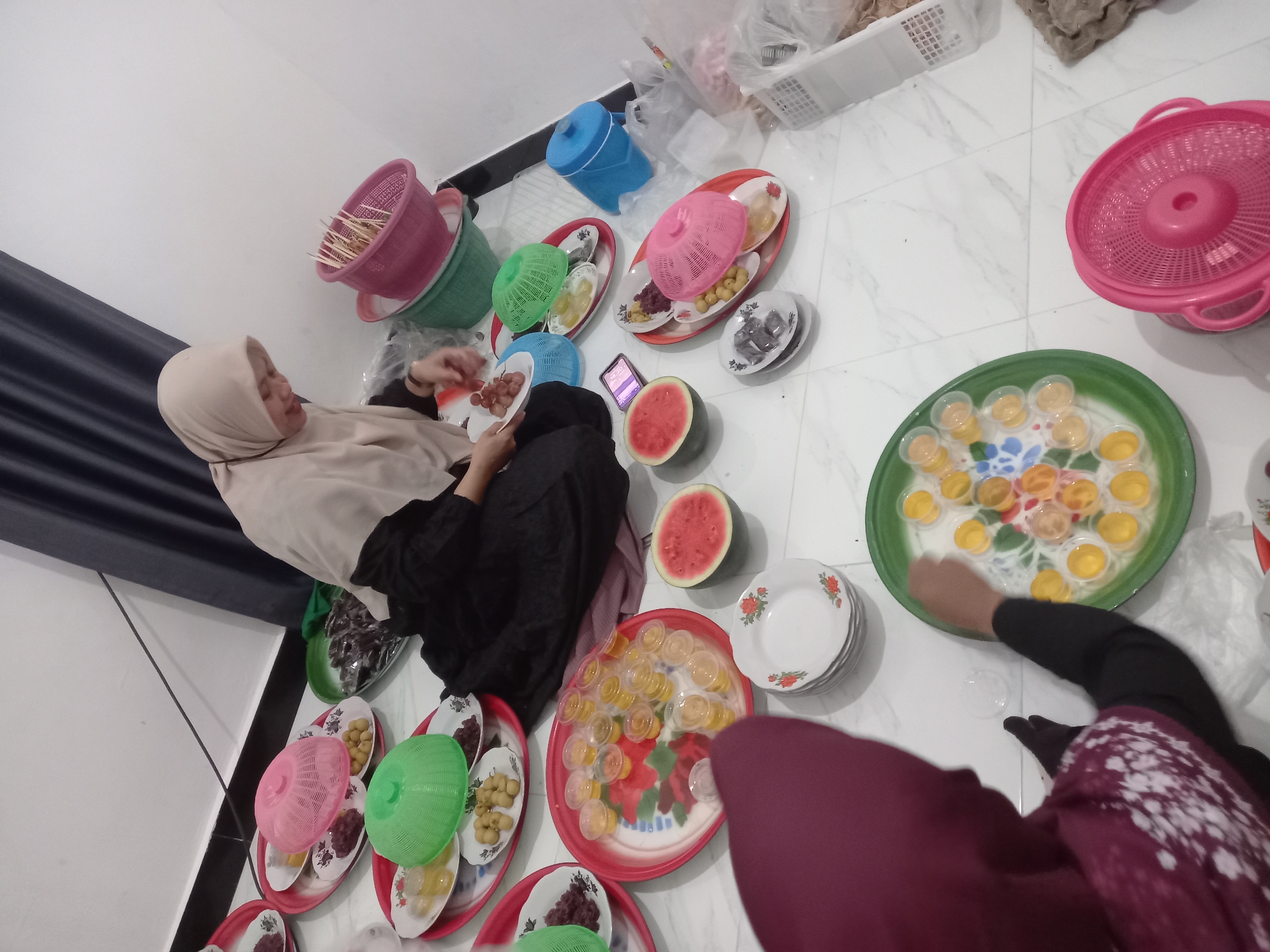 In Tempos, Indonesia, two individuals, presumably women, are preparing food on a white tiled floor. One woman, wearing a light-colored hijab and a black dress, is seated and appears to be arranging food items on a plate. In front of her, various platters hold what seem to be grapes, olives, and small, yellow, jelly-like desserts. Several colorful plastic baskets, pink, green, and blue, are scattered around. The second individual, wearing a maroon hijab, is also seated and is reaching towards a green platter filled with similar jelly desserts. A cut watermelon is visible on the floor near the platters. In the background, a dark curtain hangs behind the seated woman, and plastic bags and white baskets suggest a food preparation or serving area. The scene depicts a communal food preparation activity, likely for an event or gathering.