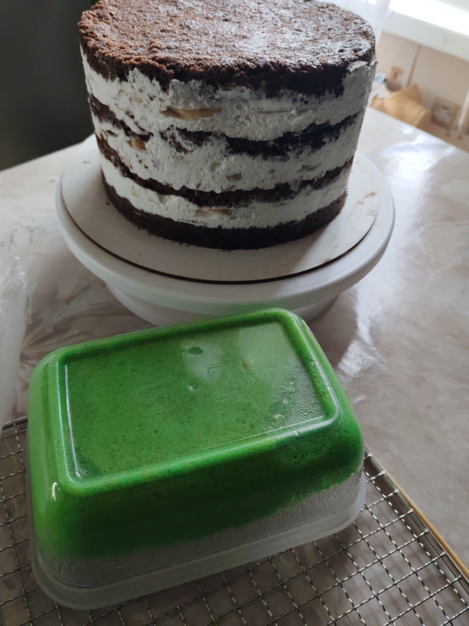 The image shows a multilayered chocolate cake with white frosting on a white cake stand. To the lower right of the cake is a green, rectangular container sitting on a wire cooling rack. The setting appears to be an indoor kitchen or food preparation area, with a marble-patterned countertop visible in the background.